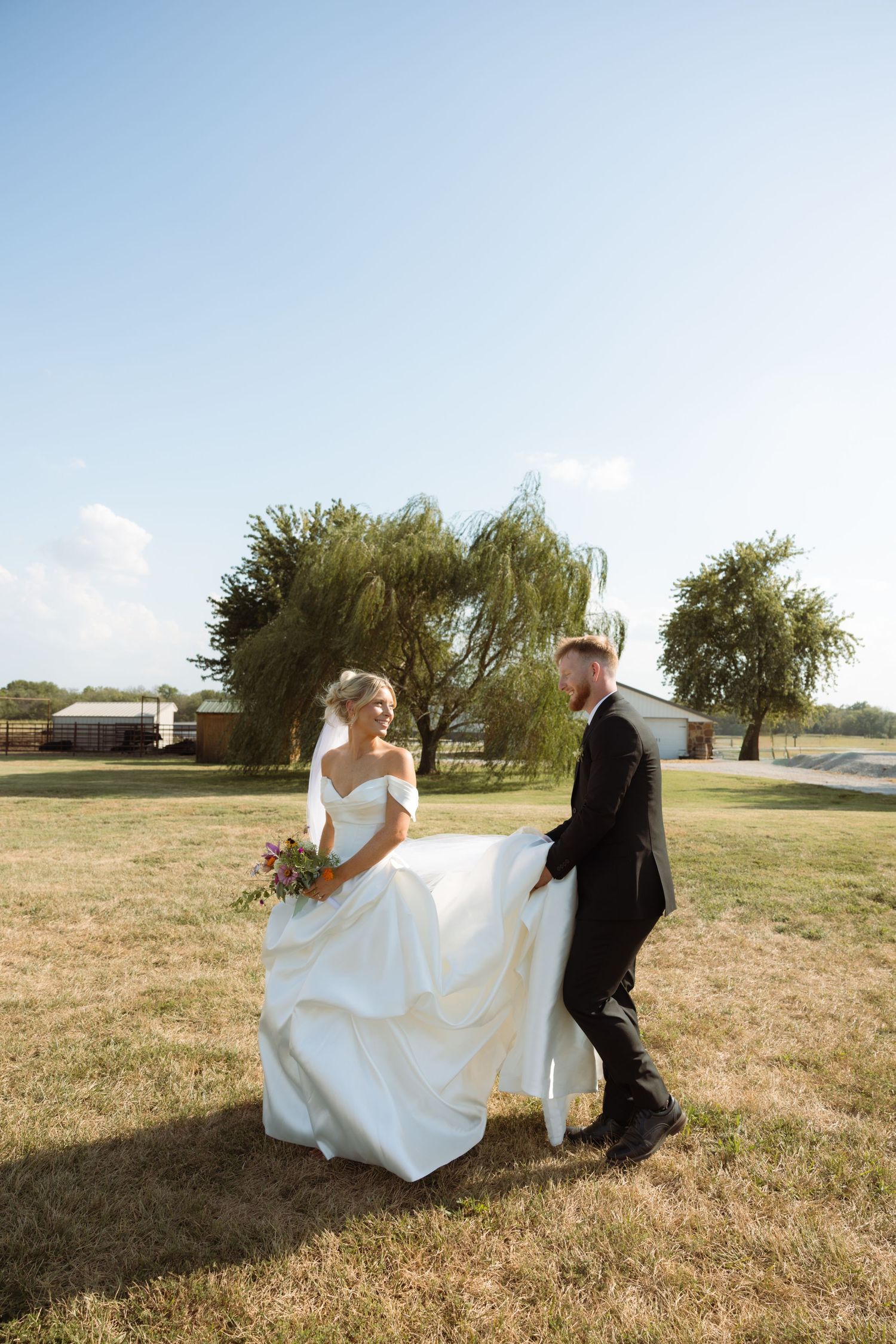 Groom helps bride carry her white wedding gown while walking across green lawn with willow trees in background.