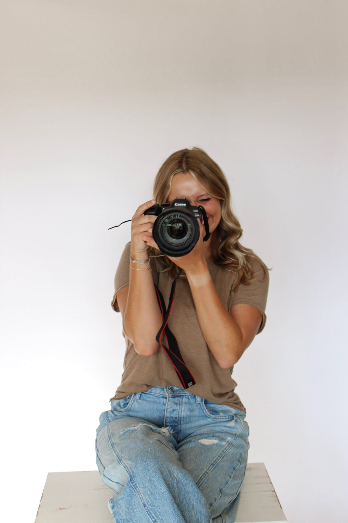 Young blonde woman photographer sitting and aiming Canon DSLR camera toward viewer.