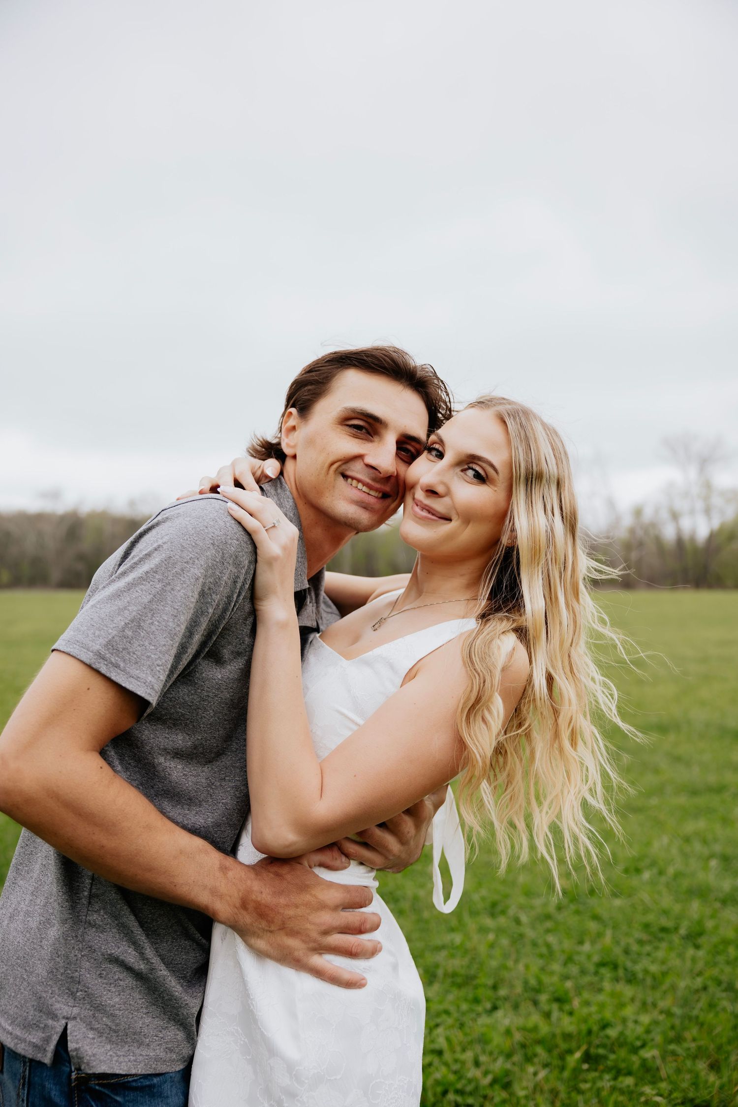 Smiling young couple embracing outdoors in green field, woman in white dress with long blonde hair.