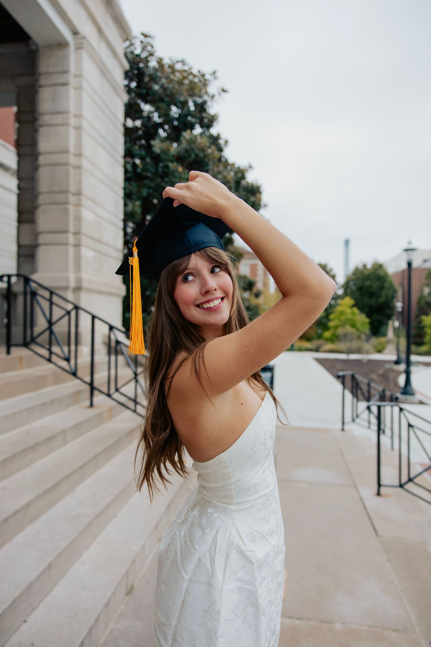 Smiling graduate in white dress adjusting black cap with gold tassel.