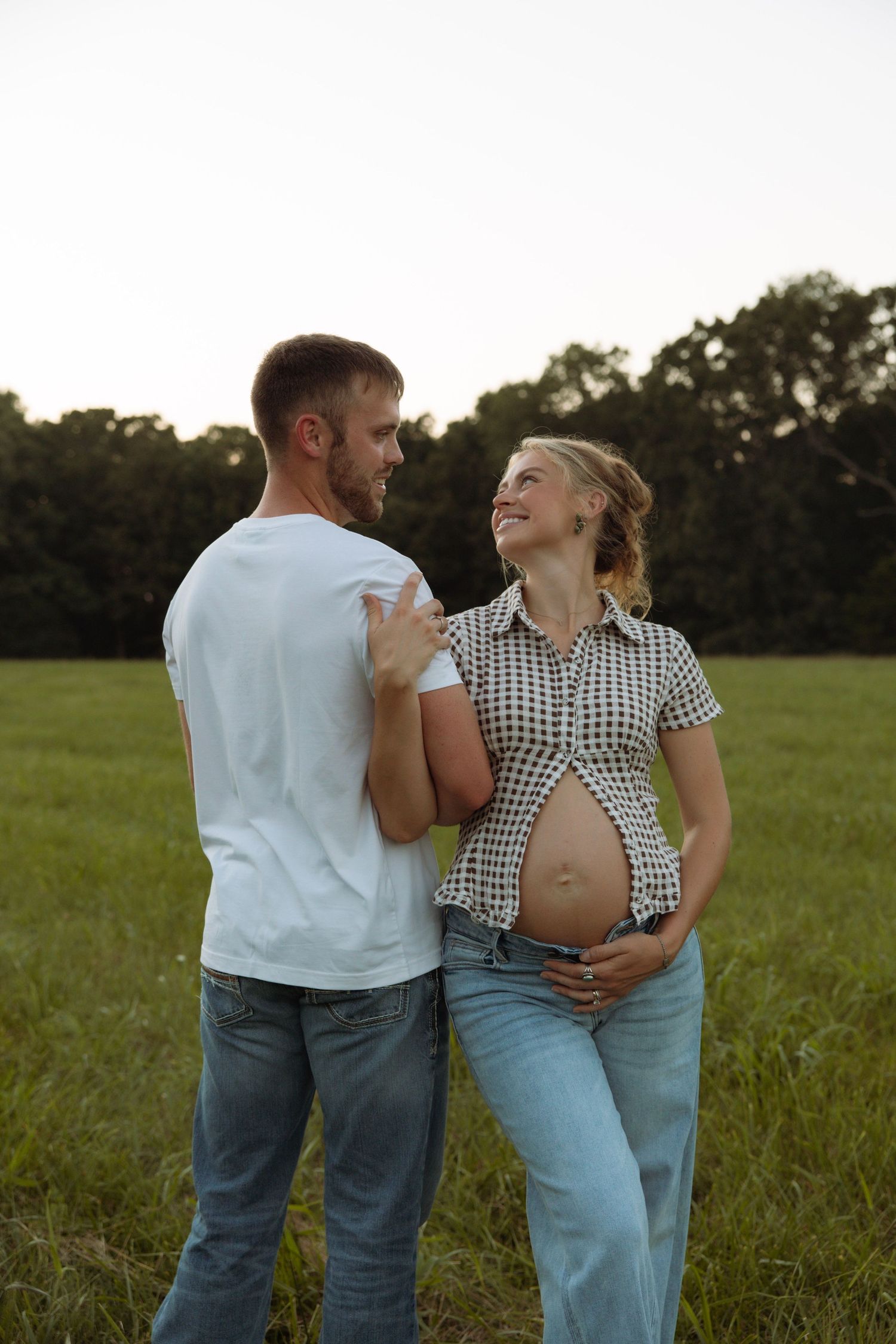 Happy pregnant couple standing in a green field at sunset, woman cradling baby bump while smiling at partner.