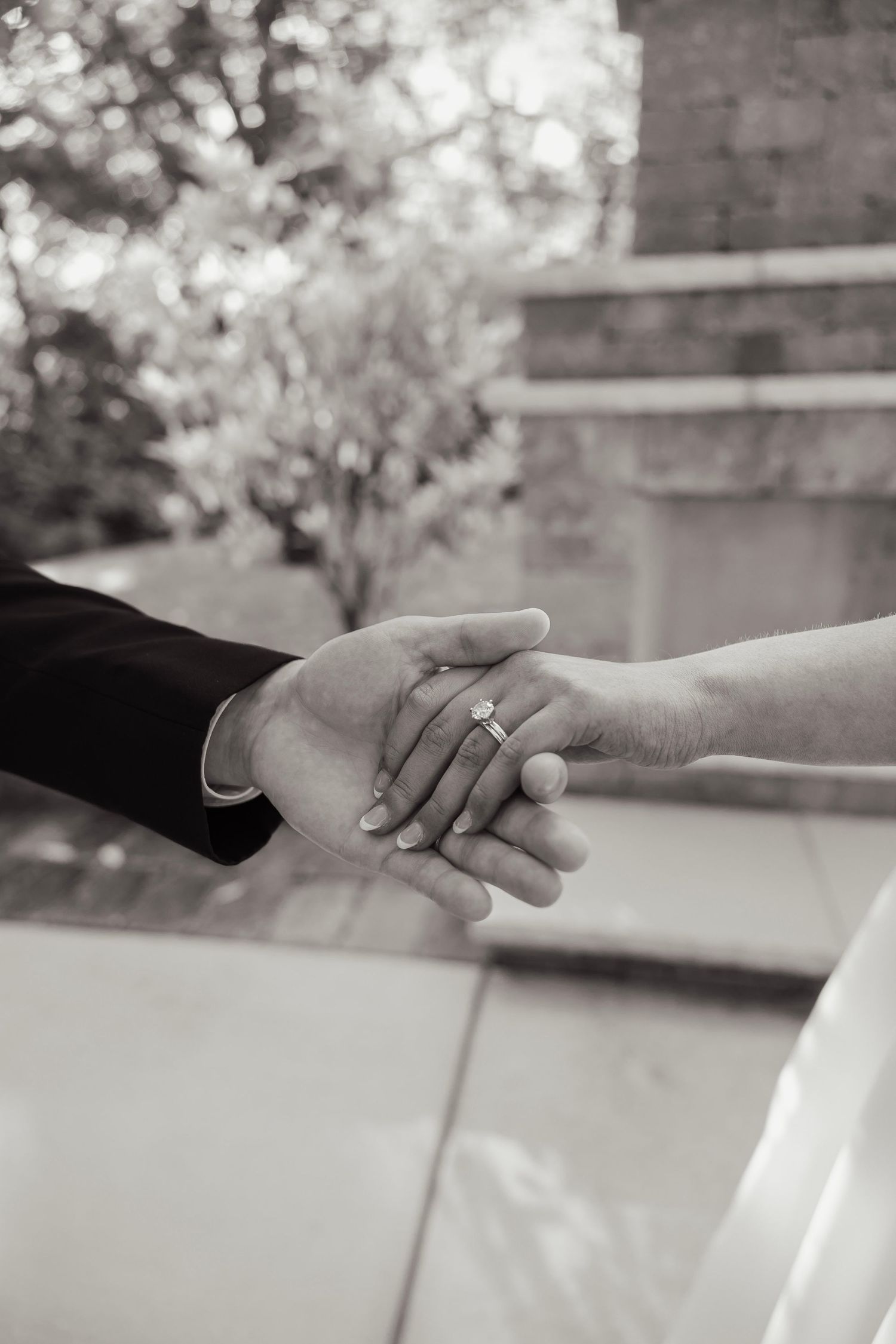 Black and white photo of a couple holding hands, showcasing a diamond engagement ring, with soft bokeh background.