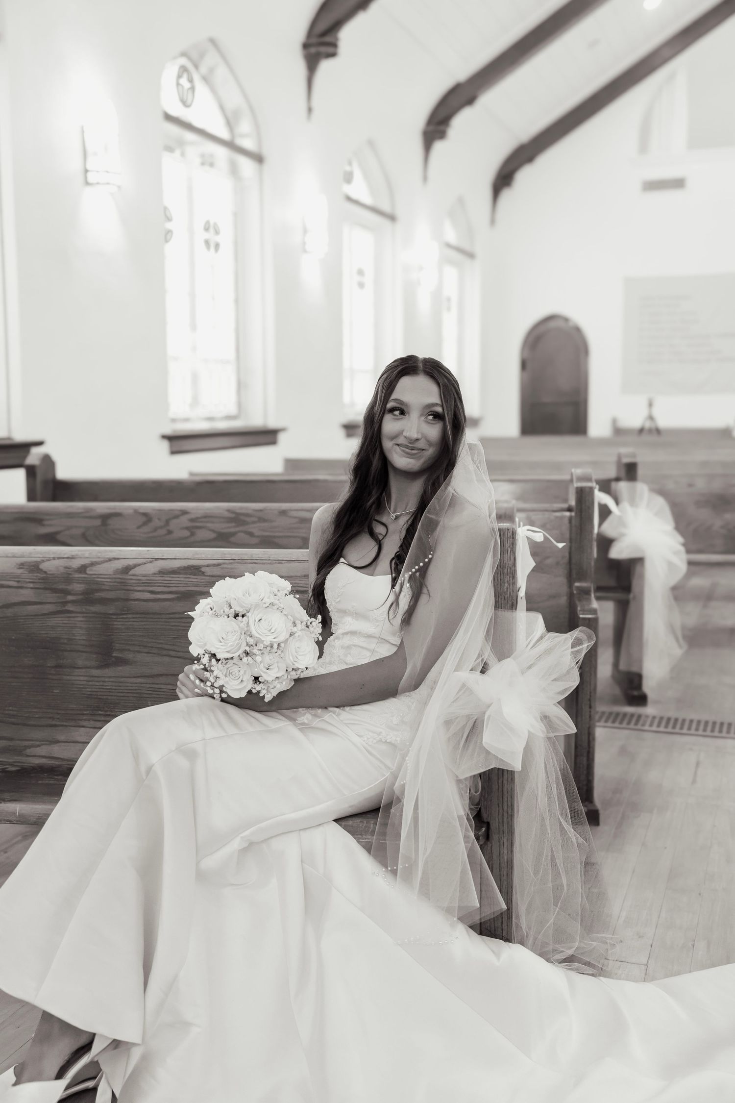 Black and white portrait of a bride in a white gown sitting in a church pew holding a bouquet.