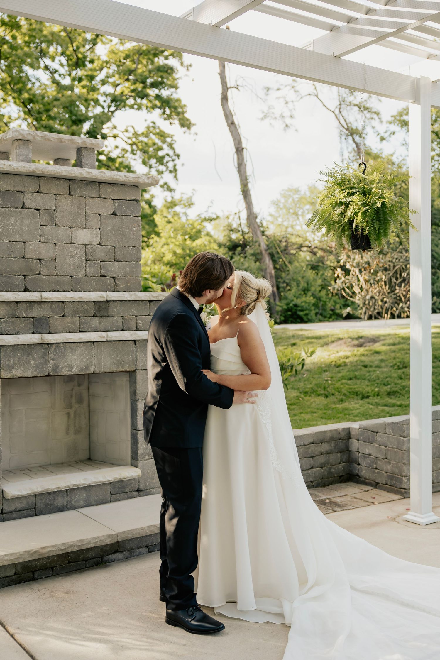 Bride and groom share first kiss under white pergola beside stone fireplace in lush garden wedding ceremony.
