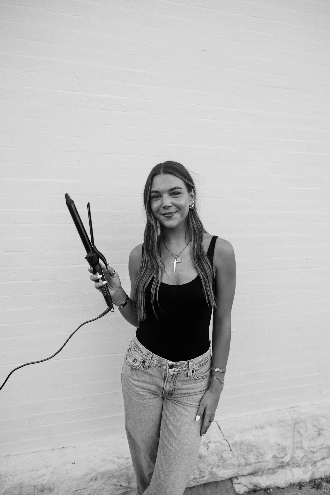 A black and white portrait of a smiling young woman holding a curling iron against a white brick wall.
