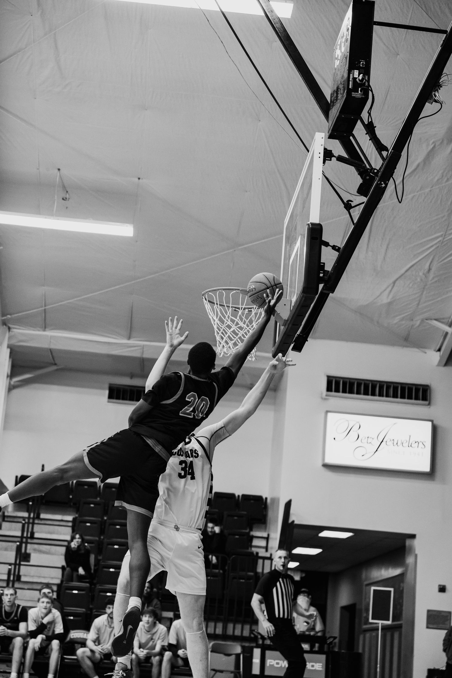 Black and white photo of basketball player number 20 attempting a layup over defender number 34 indoors.