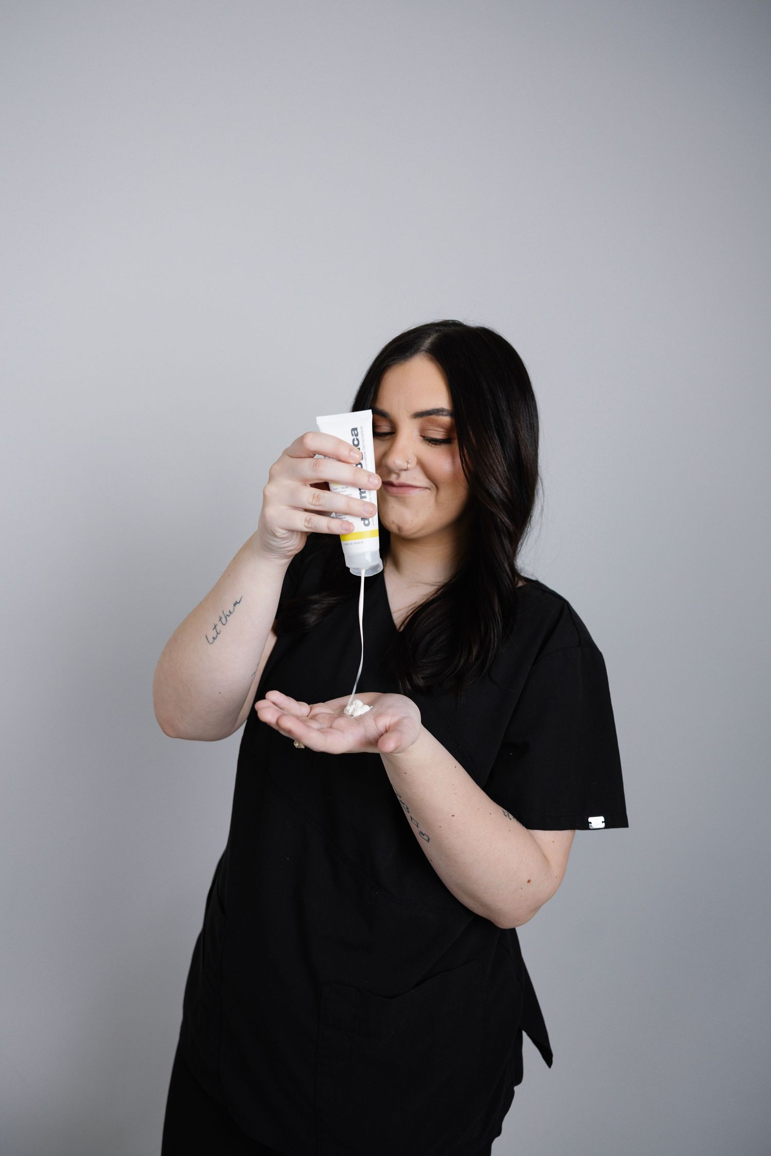 Female esthetician in black scrubs dispensing white skincare product into her palm.