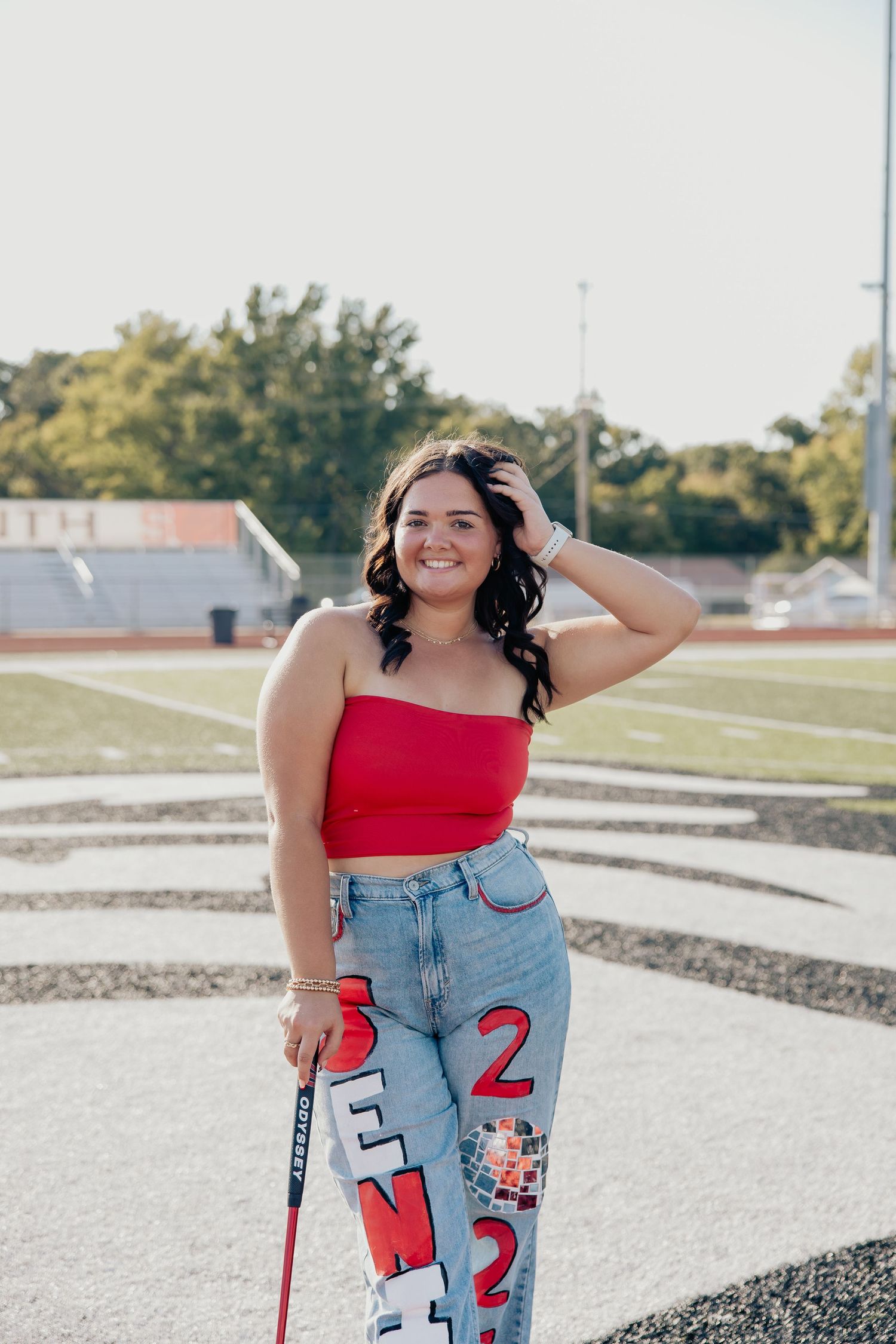 Smiling young woman in red crop top and senior jeans holding a field hockey stick on a track.