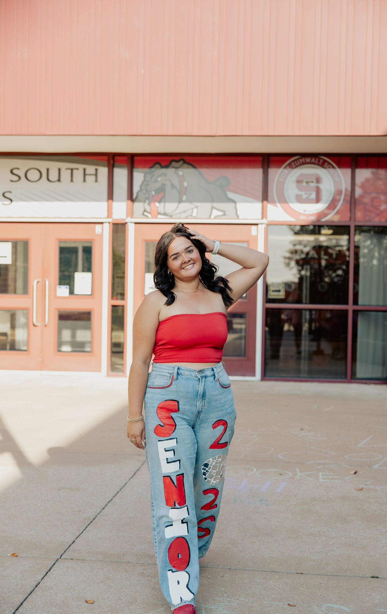Senior girl in red tube top and painted senior jeans posing in front of South high school entrance.