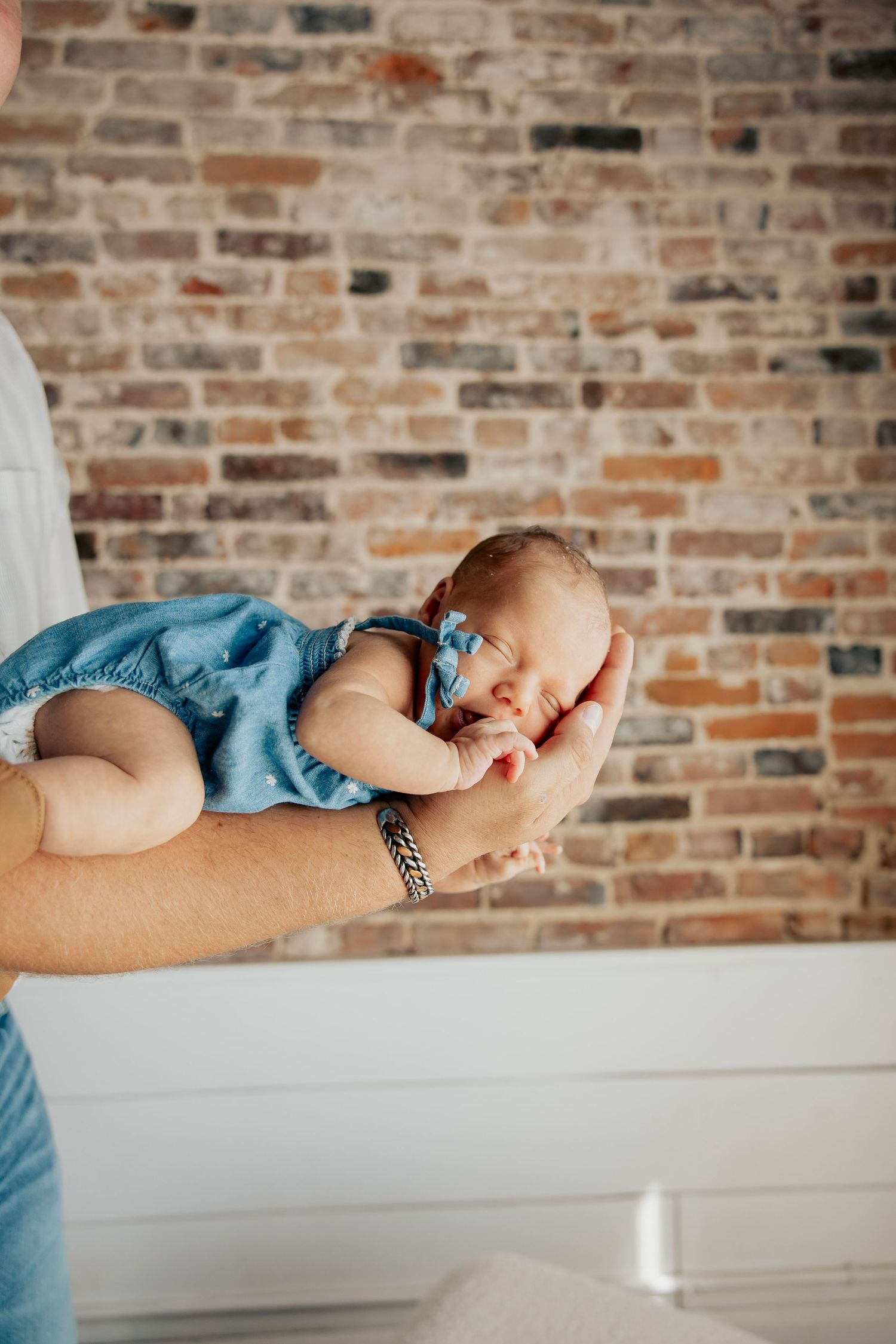 A parent gently holds a sleeping newborn baby girl in a blue romper and bow headband against a rustic brick wall backdrop.