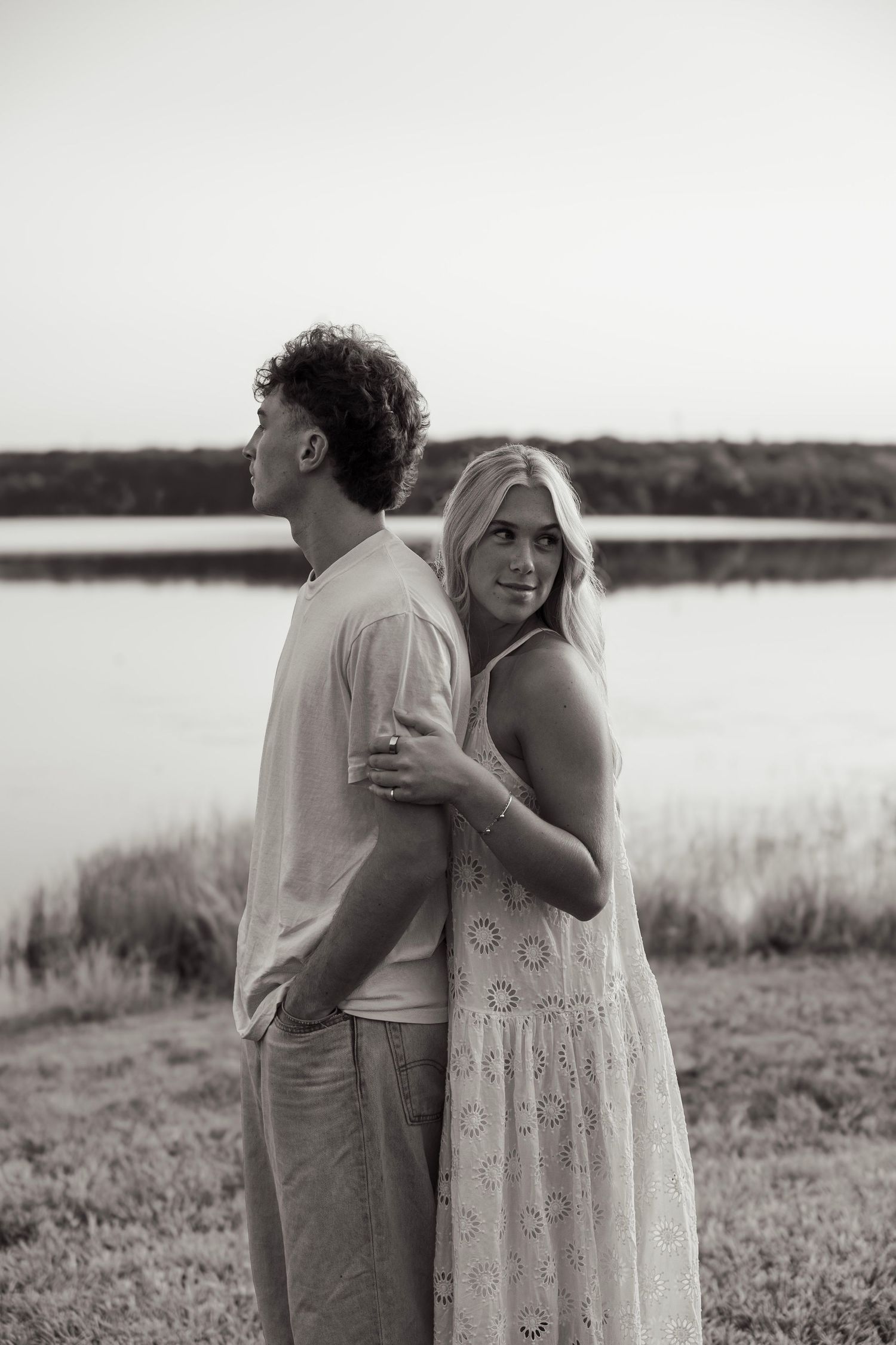 Black and white photo of a couple embracing near a lake, woman in lace dress looks at camera.