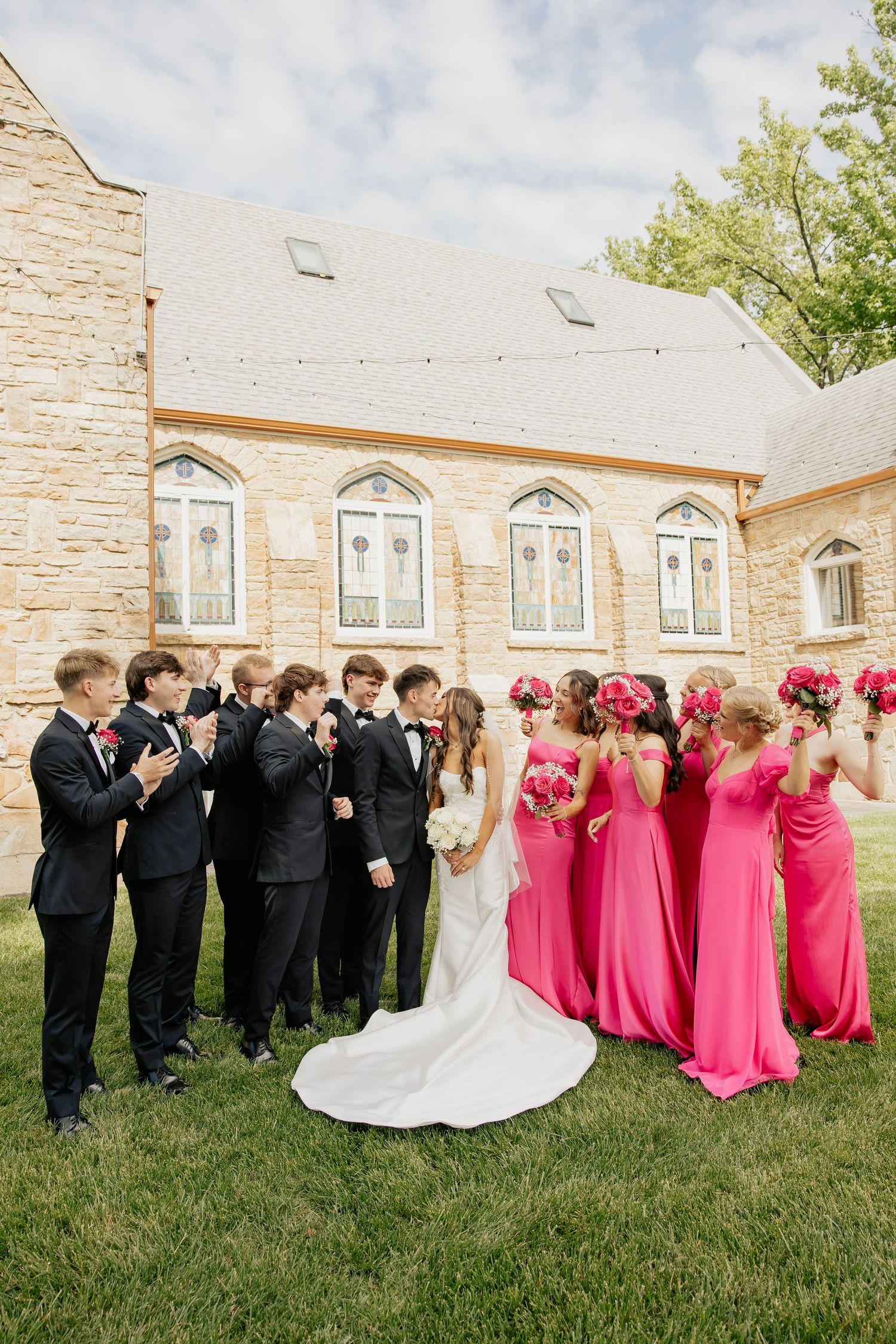 Wedding party in black tuxedos and pink gowns celebrating outside stone church.