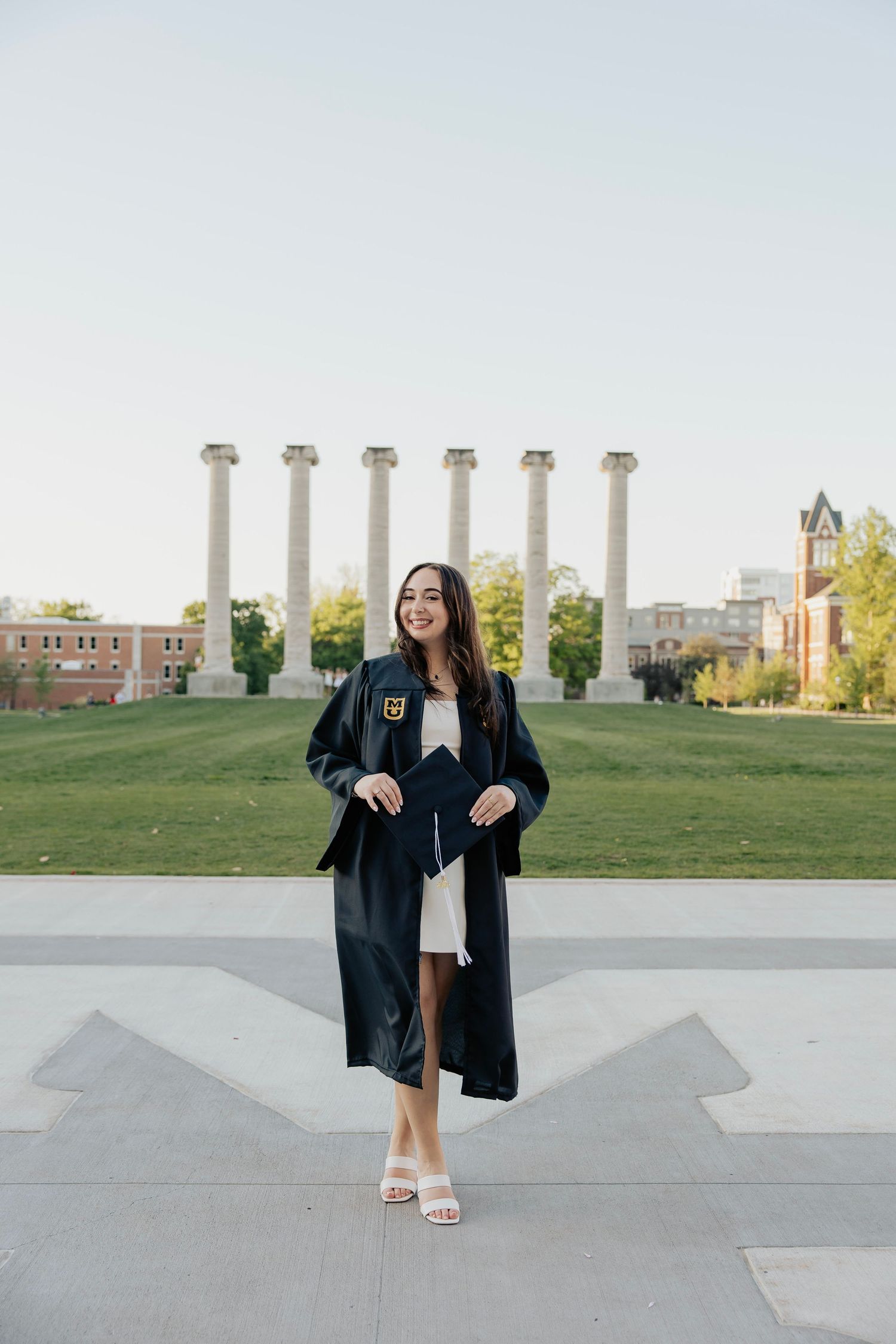 Smiling female graduate in black gown holding cap, standing before iconic university columns on campus.