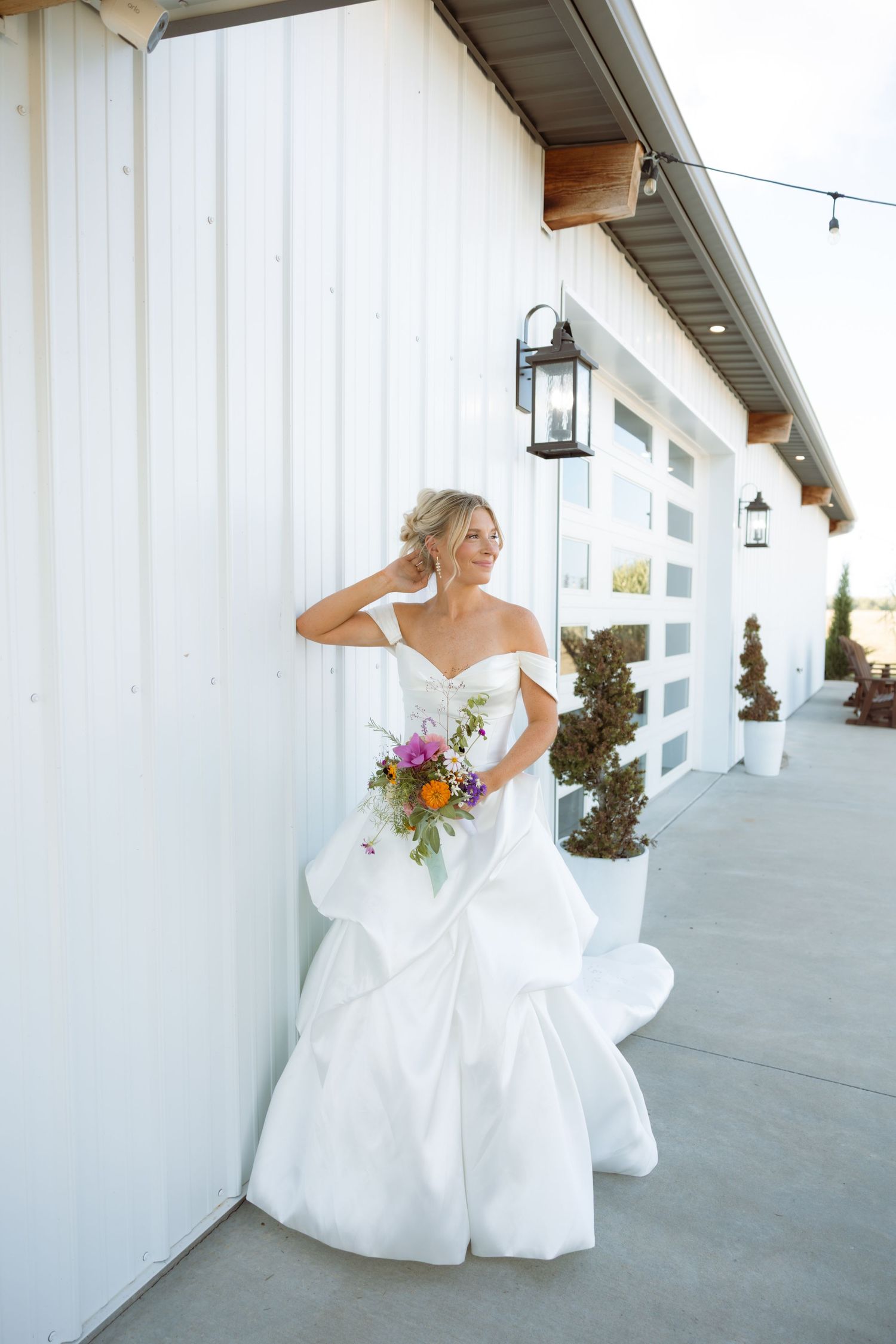 Bride in off-shoulder white mermaid gown holds colorful bouquet while posing against white barn wall with lanterns.