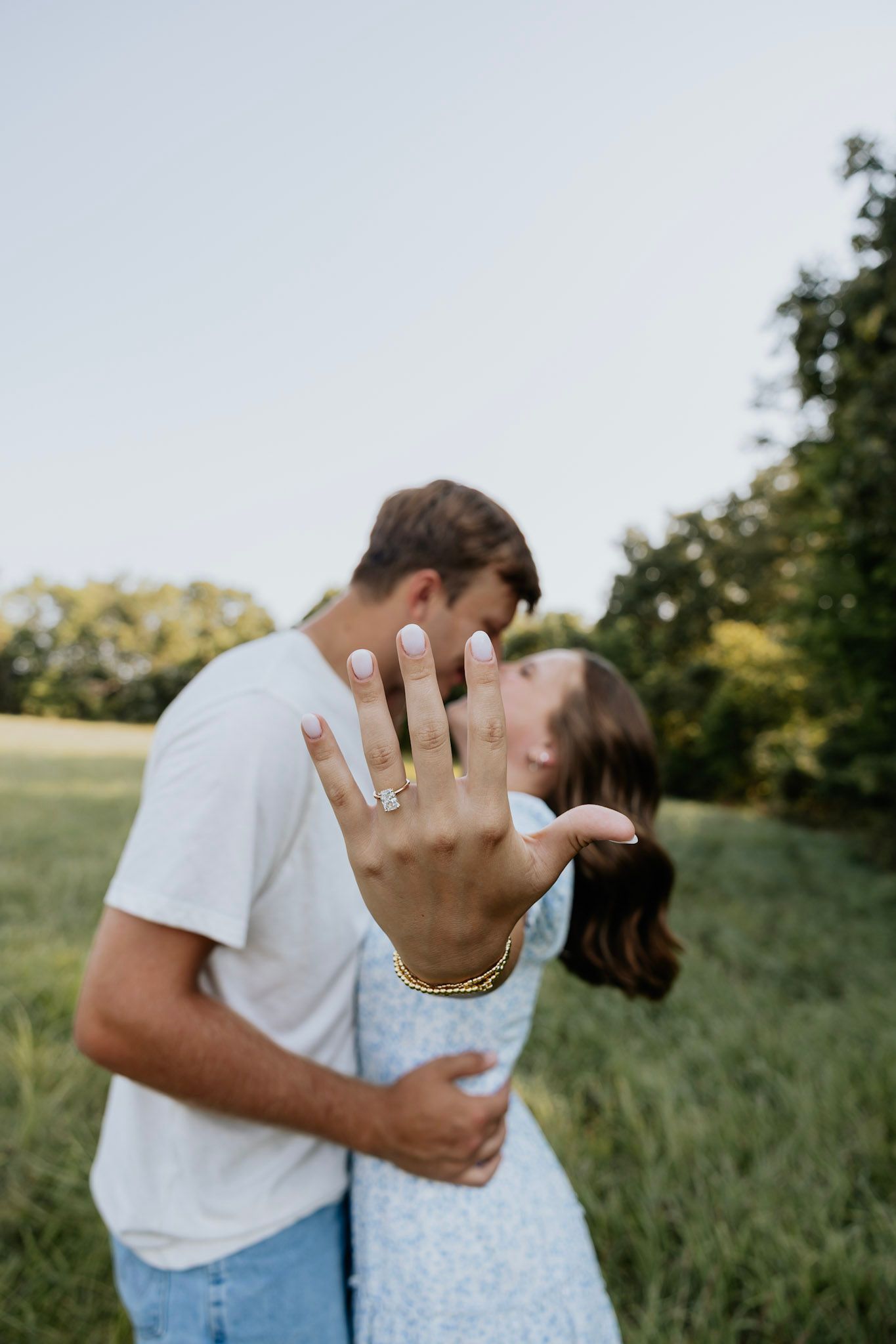 Couple kisses in green field while bride shows off engagement ring with manicured nails.