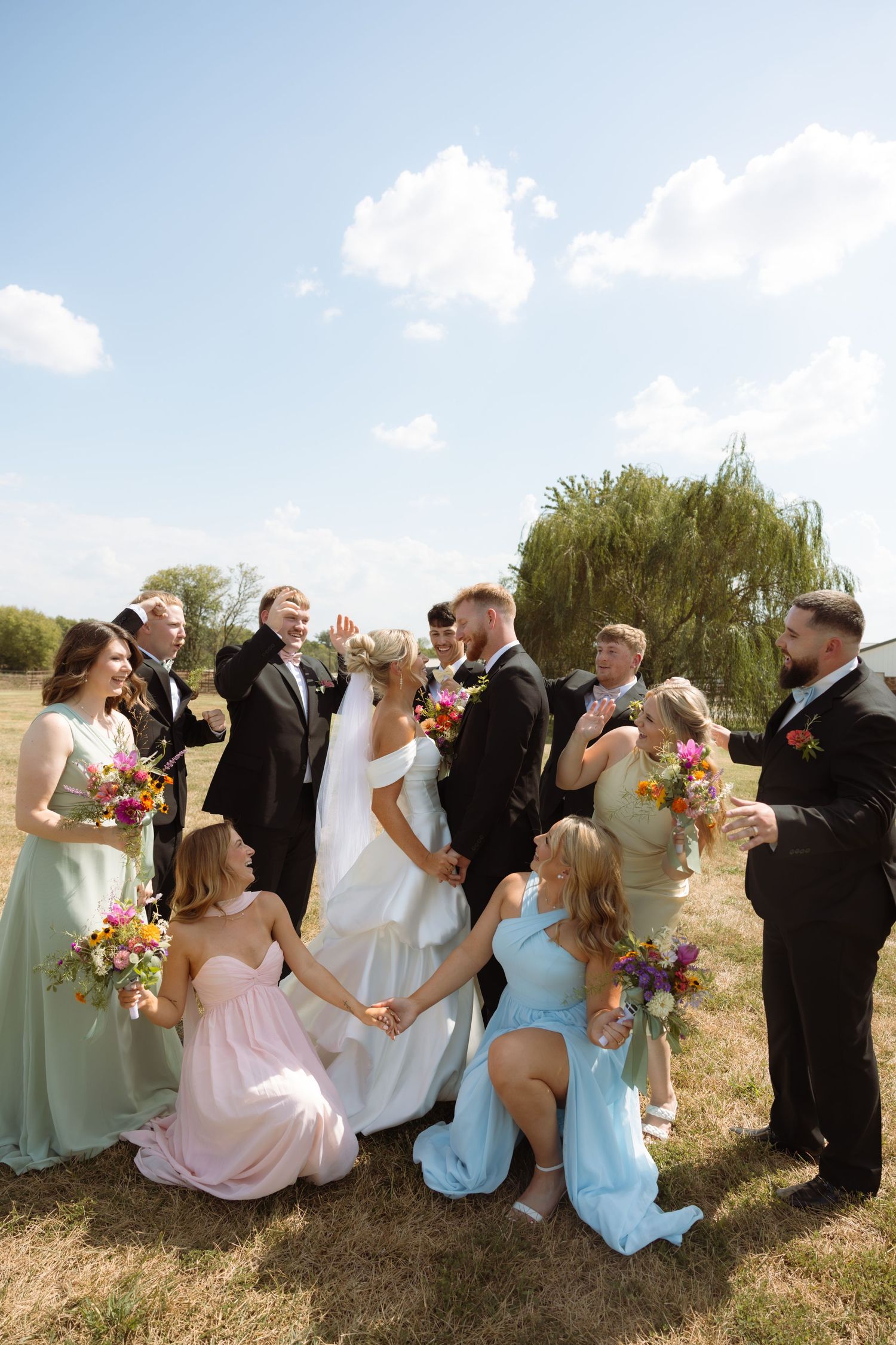 Wedding party celebrates with bride and groom outdoors, bridesmaids in colorful dresses holding bouquets on sunny day.