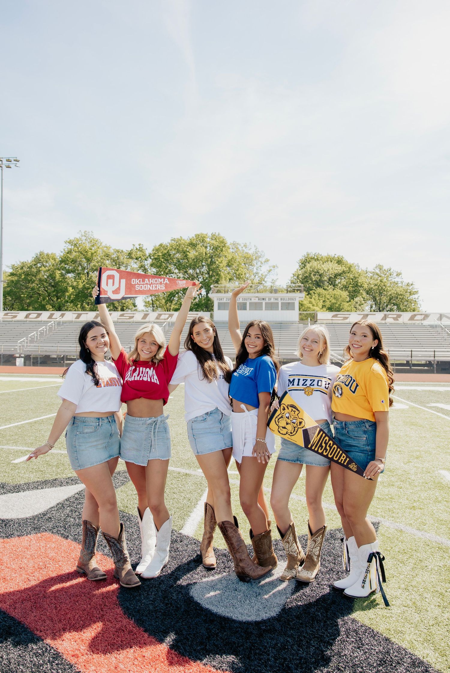 Six smiling girls in denim skirts and college shirts posing on a football field holding university pennants.