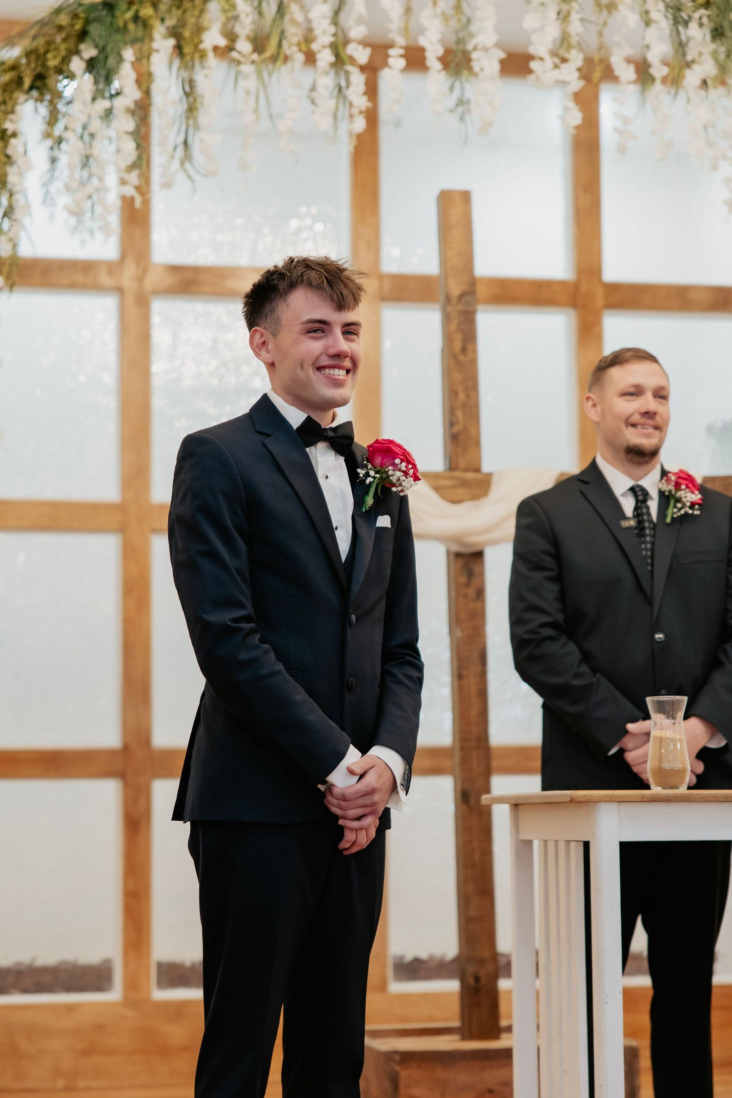 Groom in black tuxedo smiling at altar during wedding ceremony in decorated chapel.