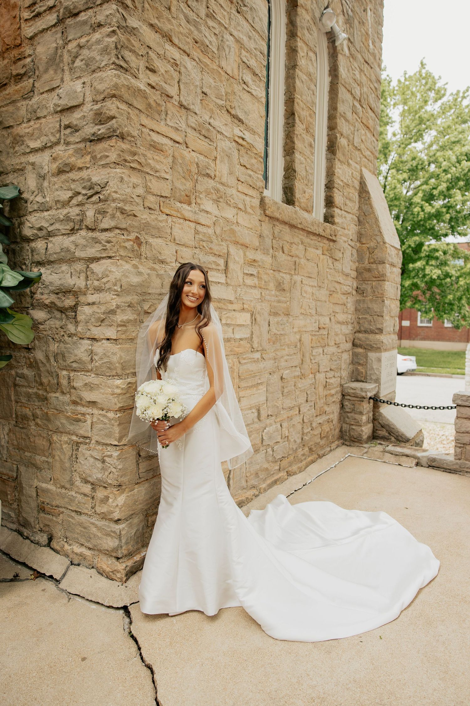 Bride in white mermaid gown with veil holding bouquet beside stone church exterior.