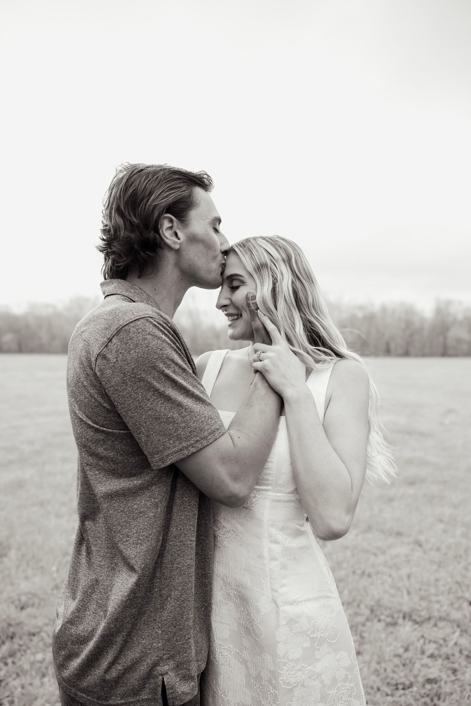 Black and white photo of a couple in a field, man kissing woman on forehead as she smiles warmly.