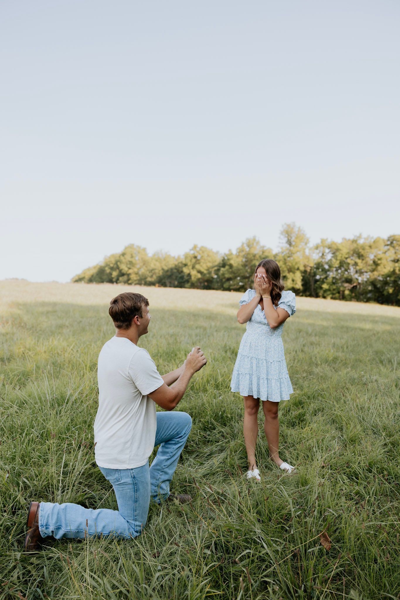 Man proposing on one knee to surprised woman in blue dress in lush green field at sunset.