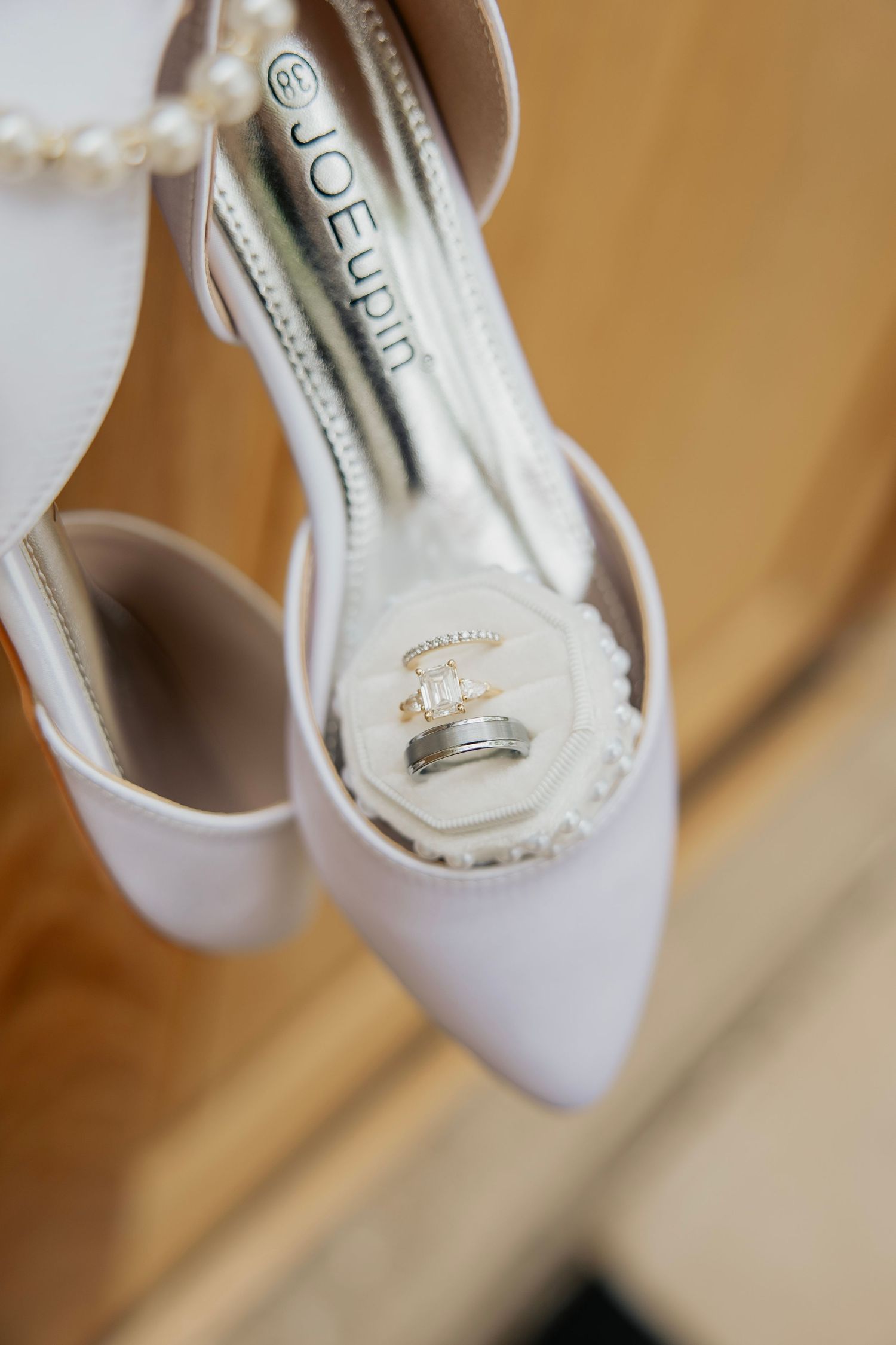 Wedding rings resting inside white pointed-toe bridal heels with pearl bracelet accent.
