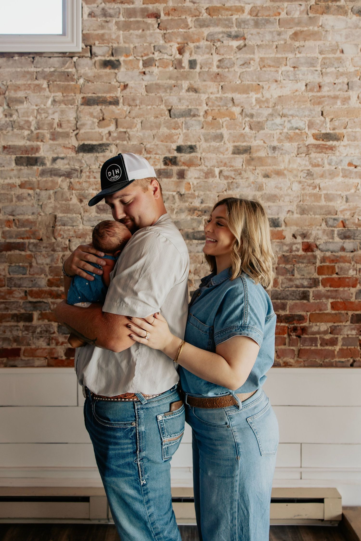 A couple in matching denim outfits stands against a brick wall, the man in a cap tenderly holding a newborn baby.