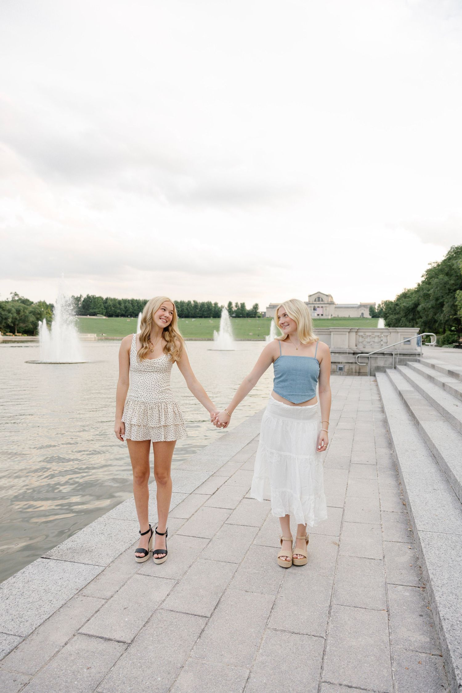 Two young blonde women holding hands and walking along a waterfront promenade with fountains in the background.