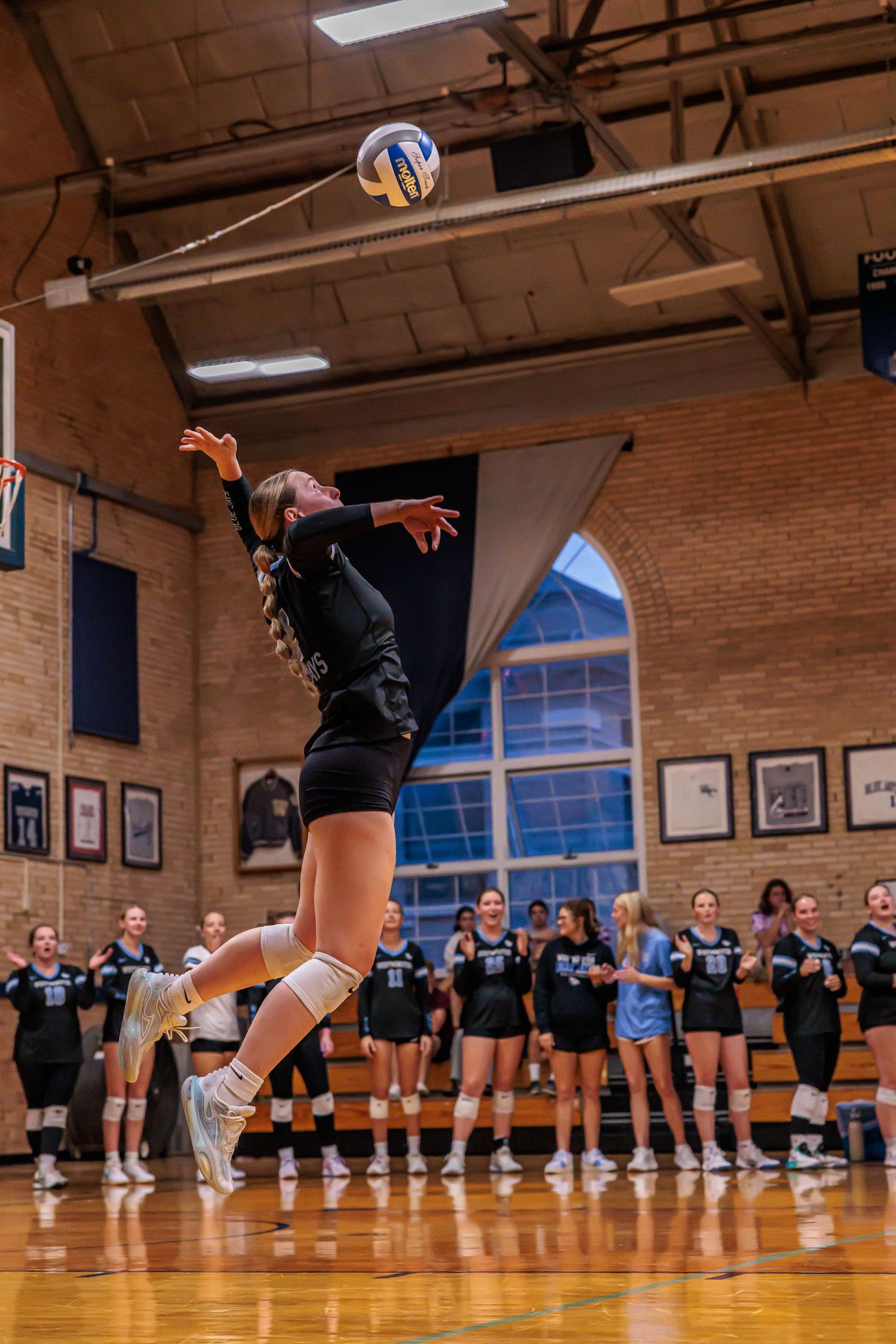 Female volleyball player in black uniform jumps to spike ball during indoor gym match with teammates watching.