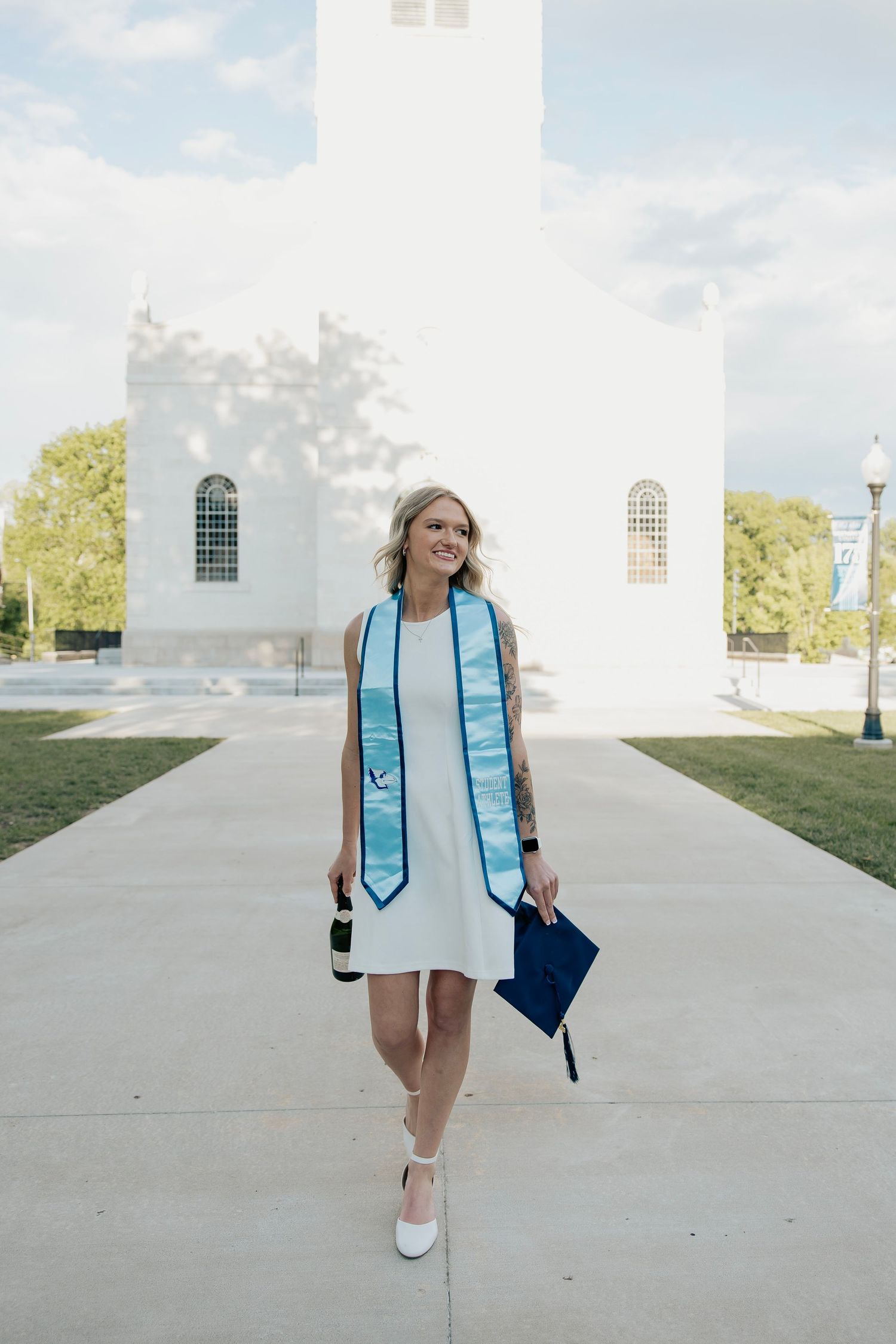 Female graduate walking in white dress with blue stole and navy cap near white building.