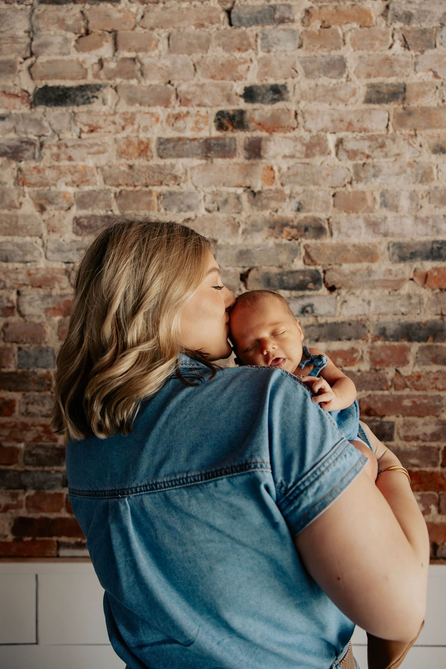 A mother in a denim shirt kisses her newborn baby against a rustic brick wall backdrop.