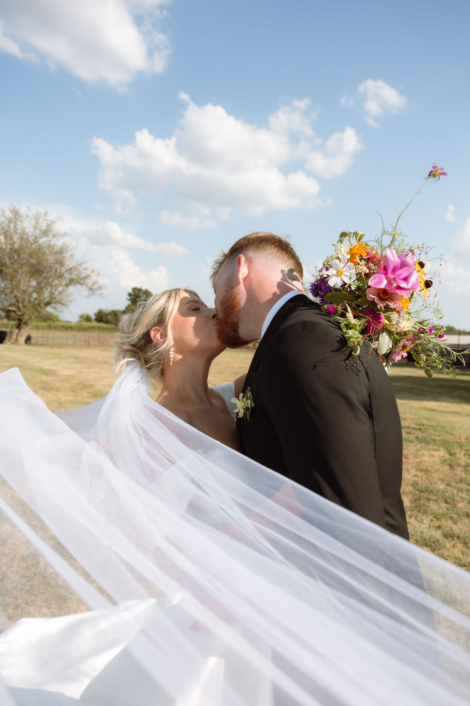 A bride and groom share a kiss outdoors, her veil flowing in the wind, while she holds a colorful wildflower bouquet.