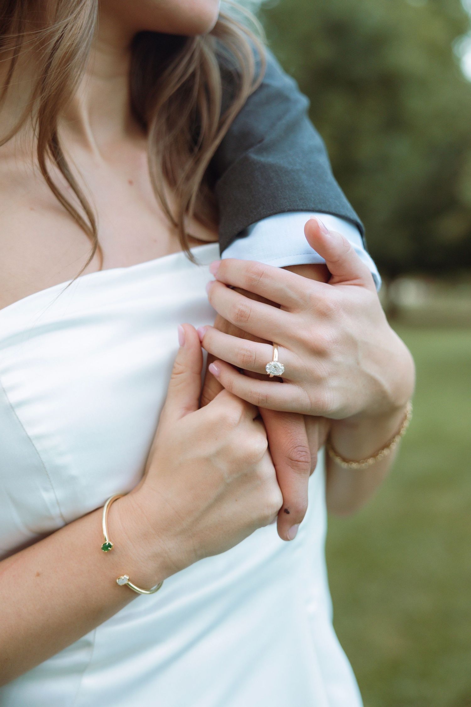 Bride in white dress showing diamond engagement ring and gold bracelet while holding groom's arm outdoors.