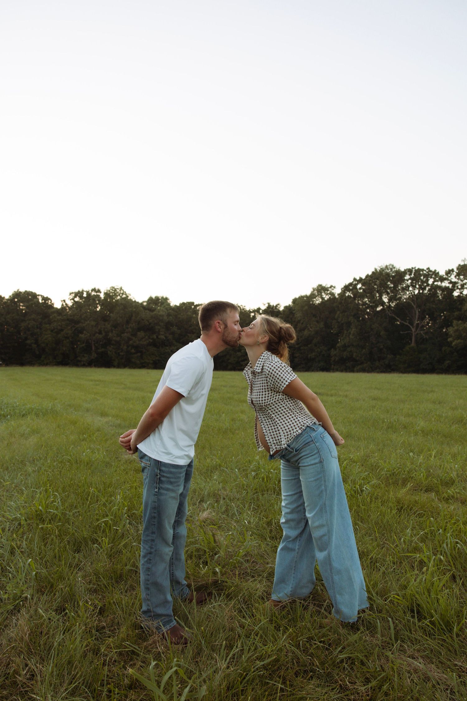 A couple sharing a romantic kiss in a lush green field with trees in the background.