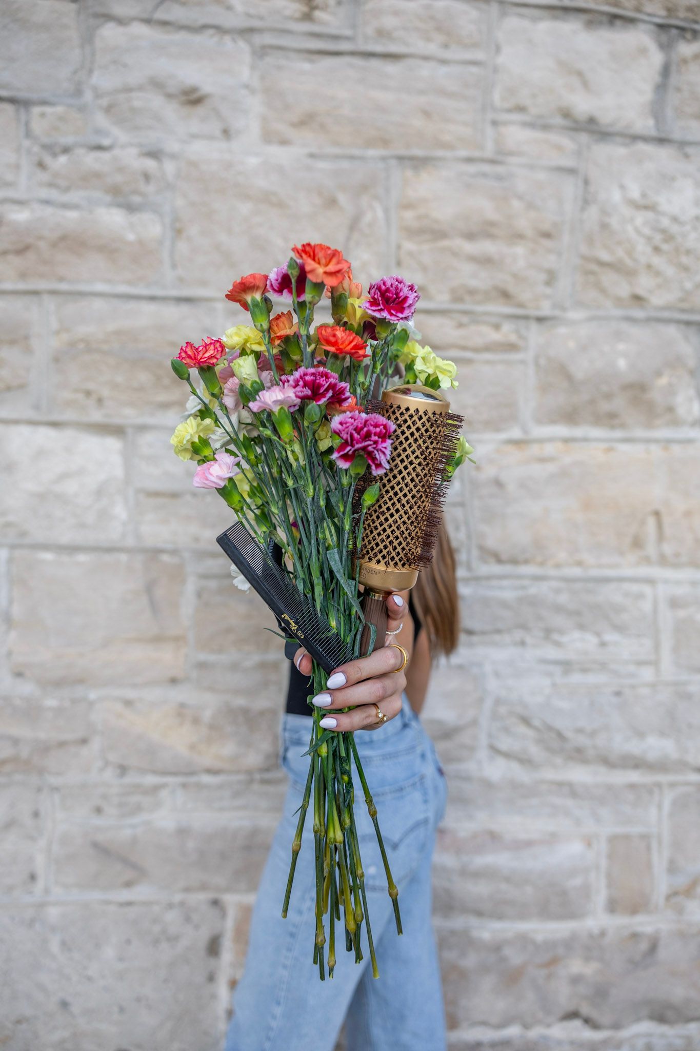 Woman extending vibrant mixed carnation bouquet with gold round brush toward camera against stone wall.
