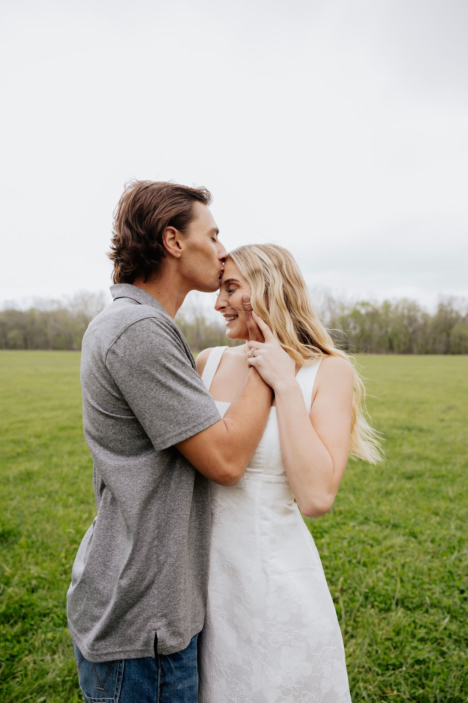 Young couple embracing in green field, man kissing womans temple while she smiles joyfully.