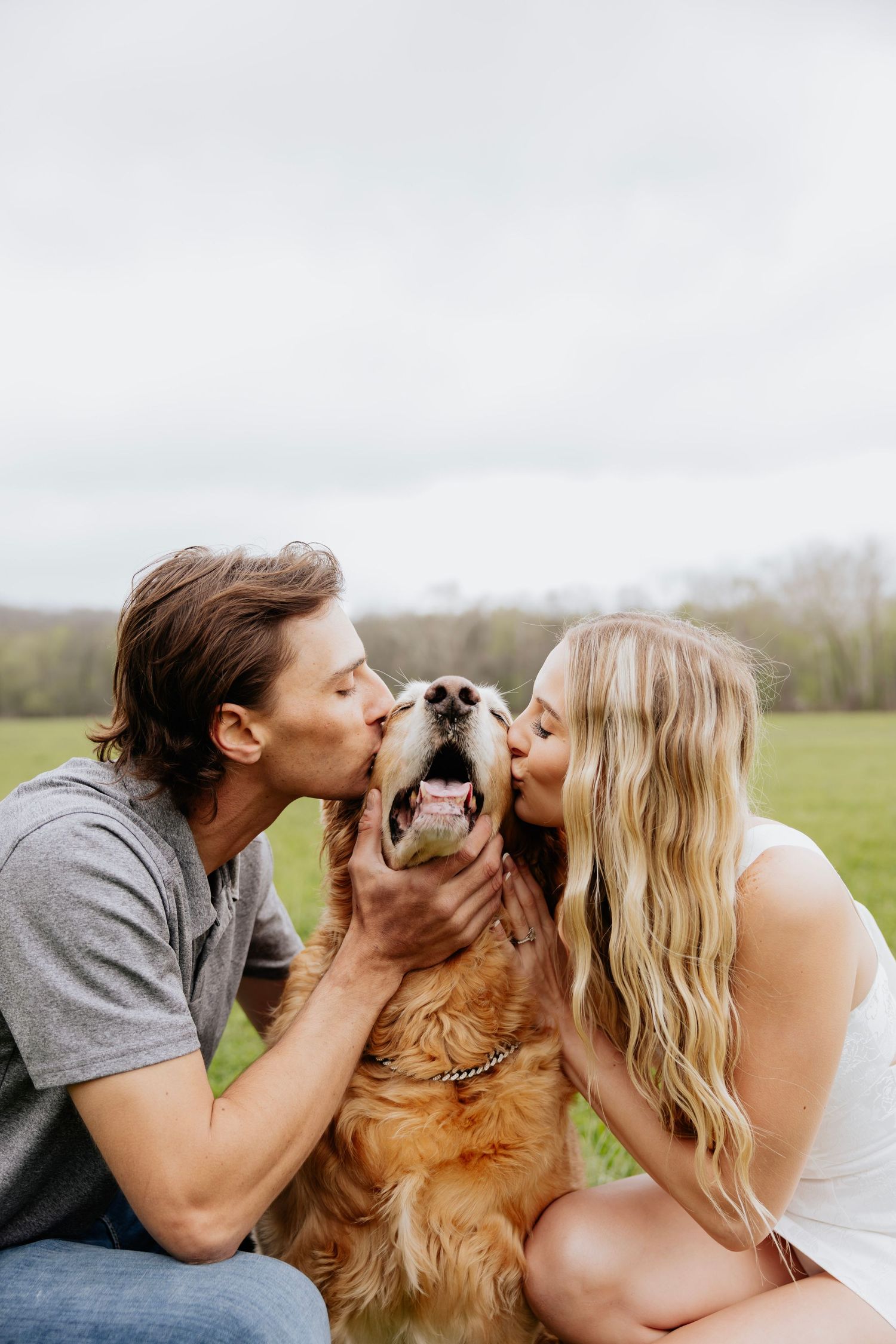 Couple kissing a happy golden retriever on both cheeks while sitting in a lush green field.