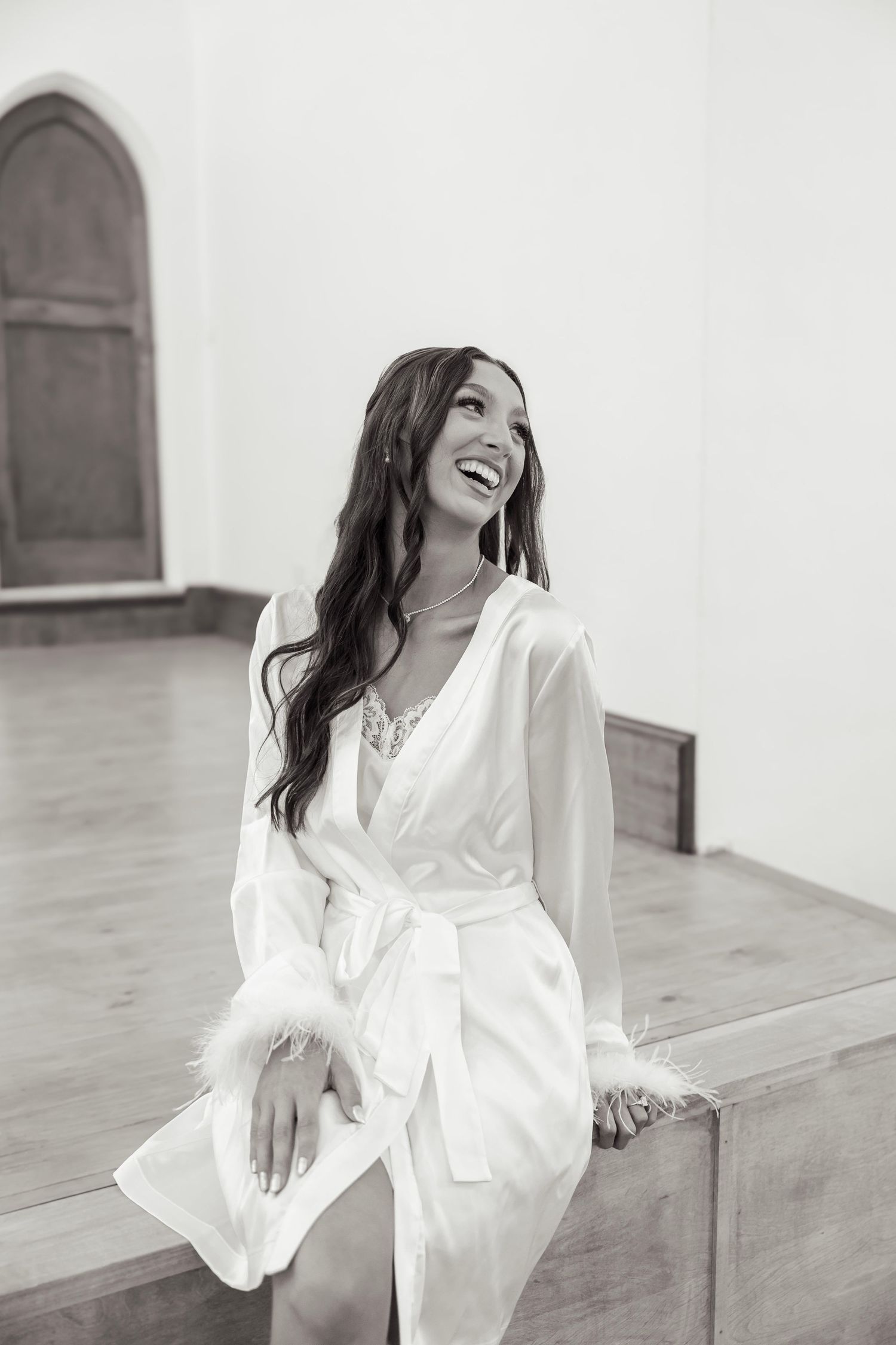 Black and white photo of a laughing bride in a white feather robe sitting on a church pew.
