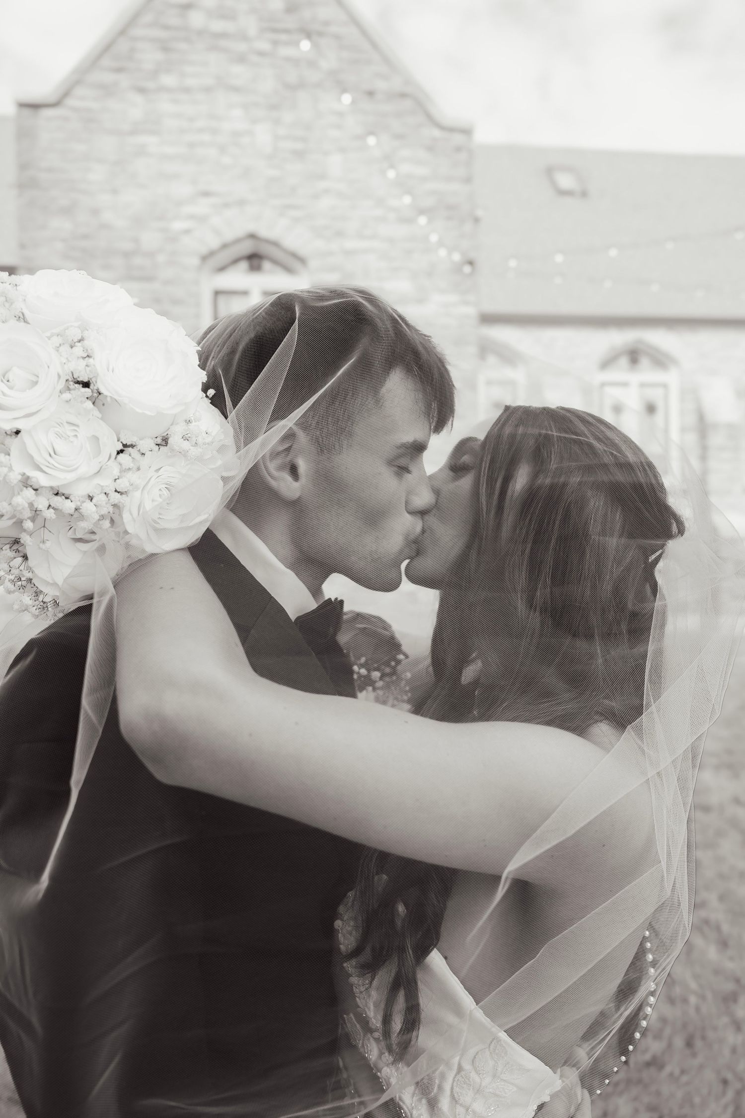 Black and white romantic photo of newlyweds kissing under bridal veil outside stone church.