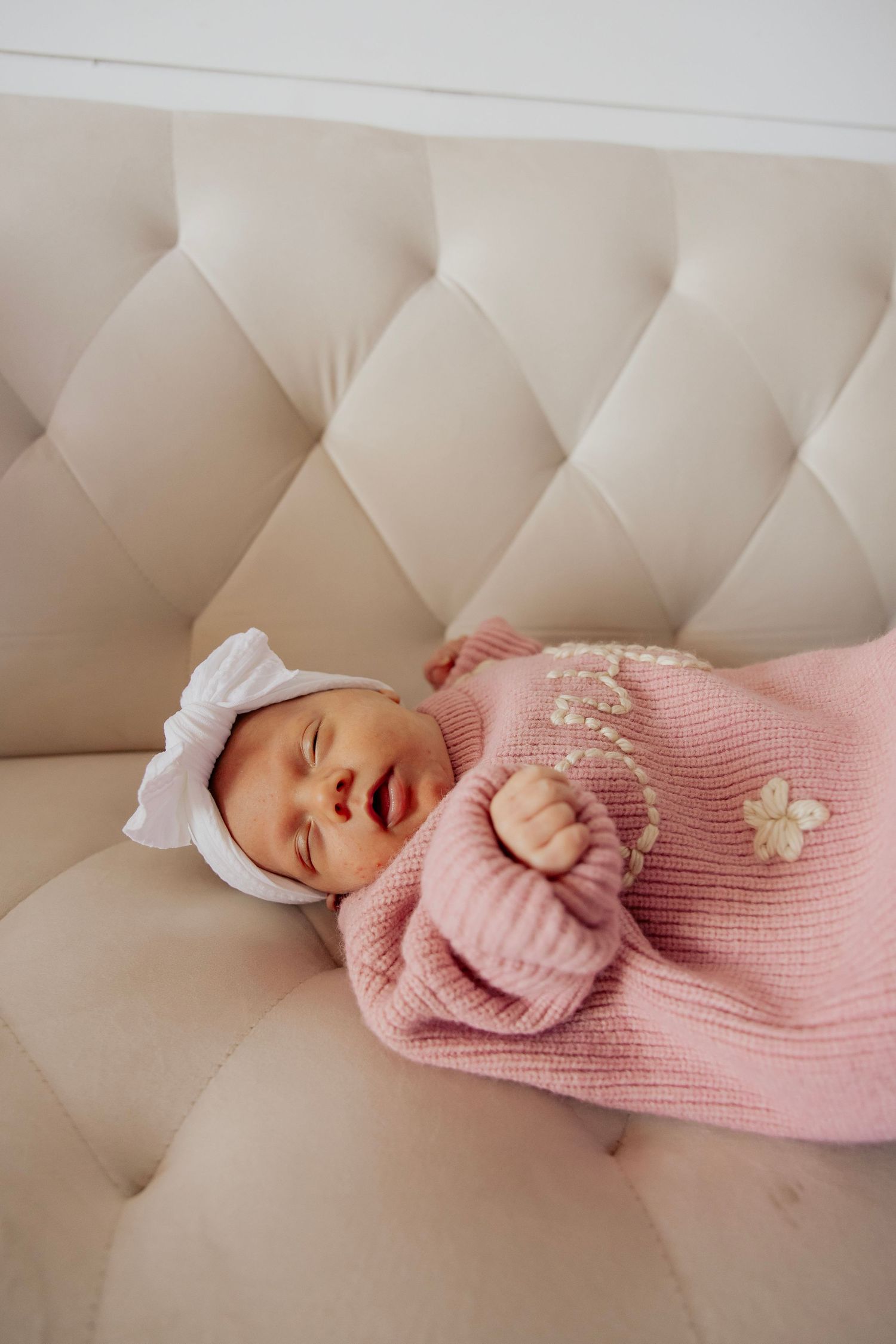 Newborn baby in pink knit outfit and white bow hat resting on a tufted beige sofa.