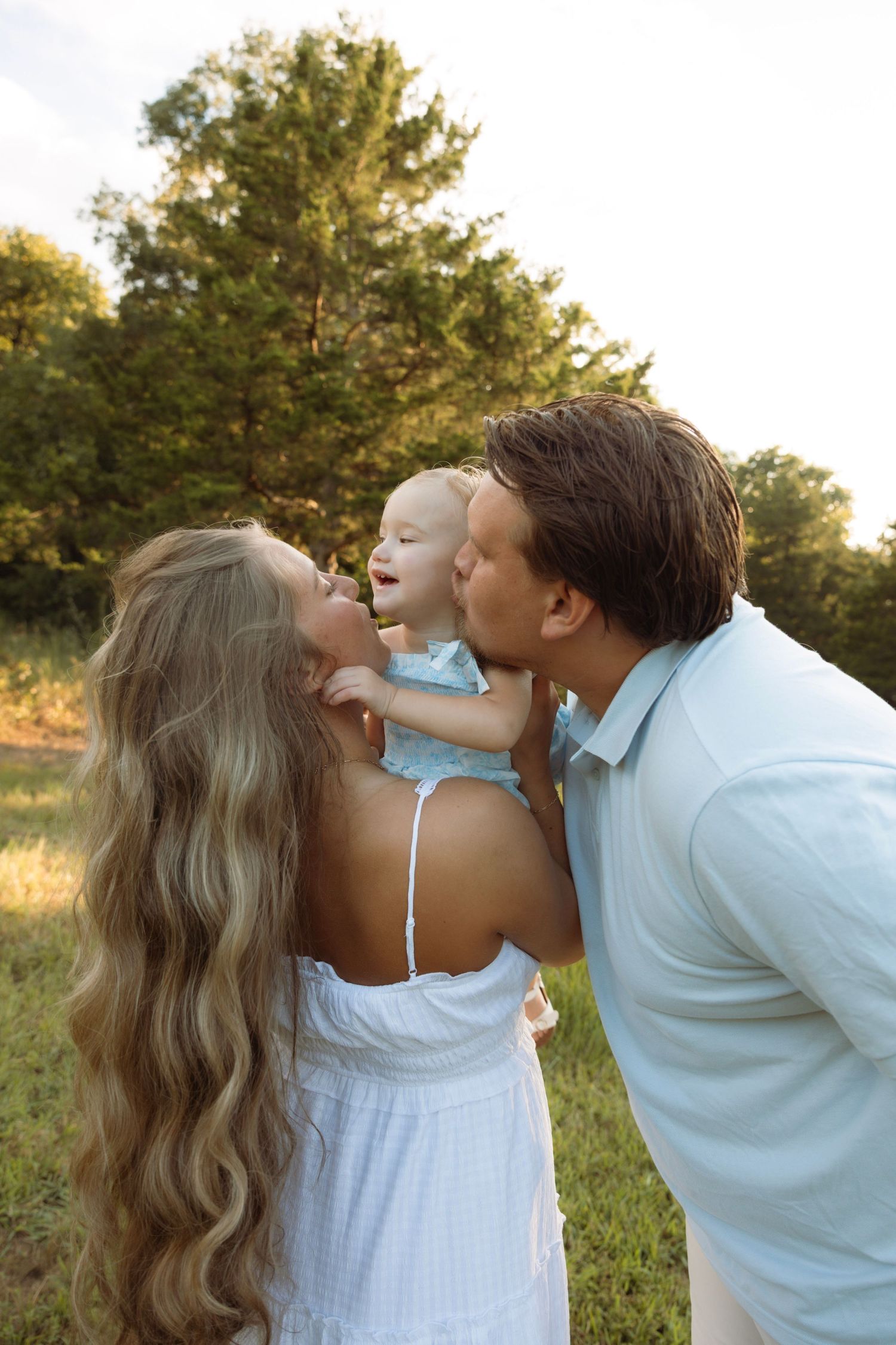 A couple in white outfits kisses their laughing baby outdoors in a sunny green field.
