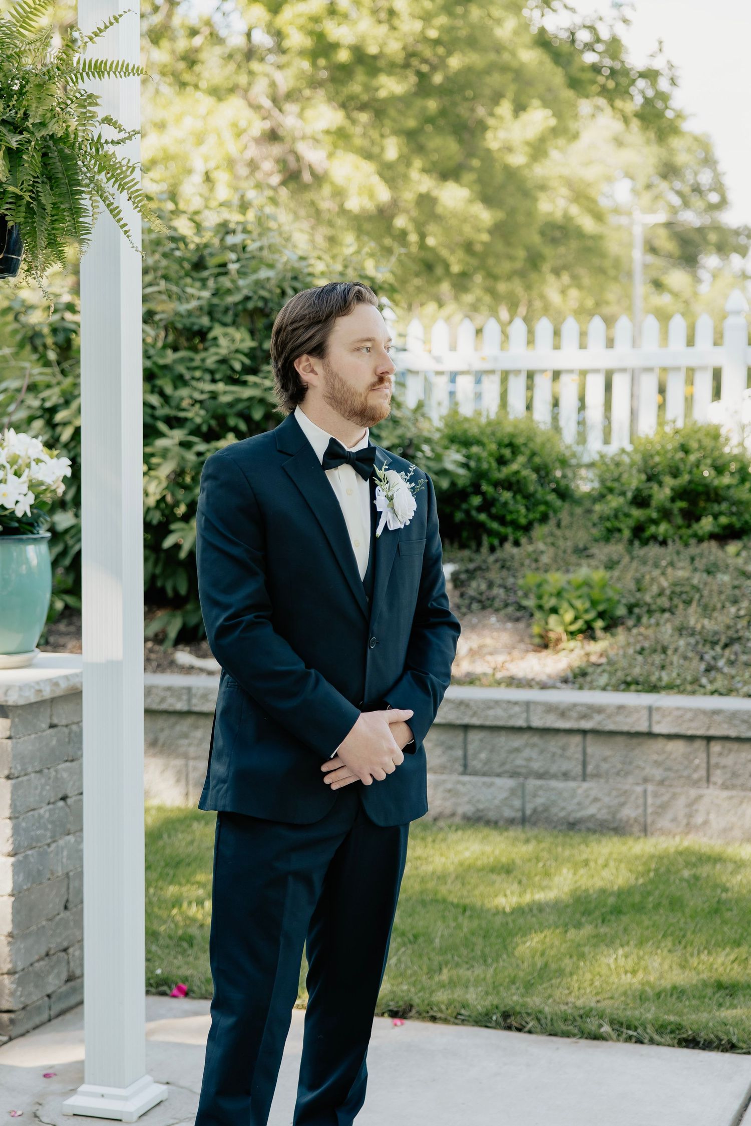 Groom in navy tuxedo with white boutonniere stands outdoors at wedding ceremony beside white fence and lush greenery.