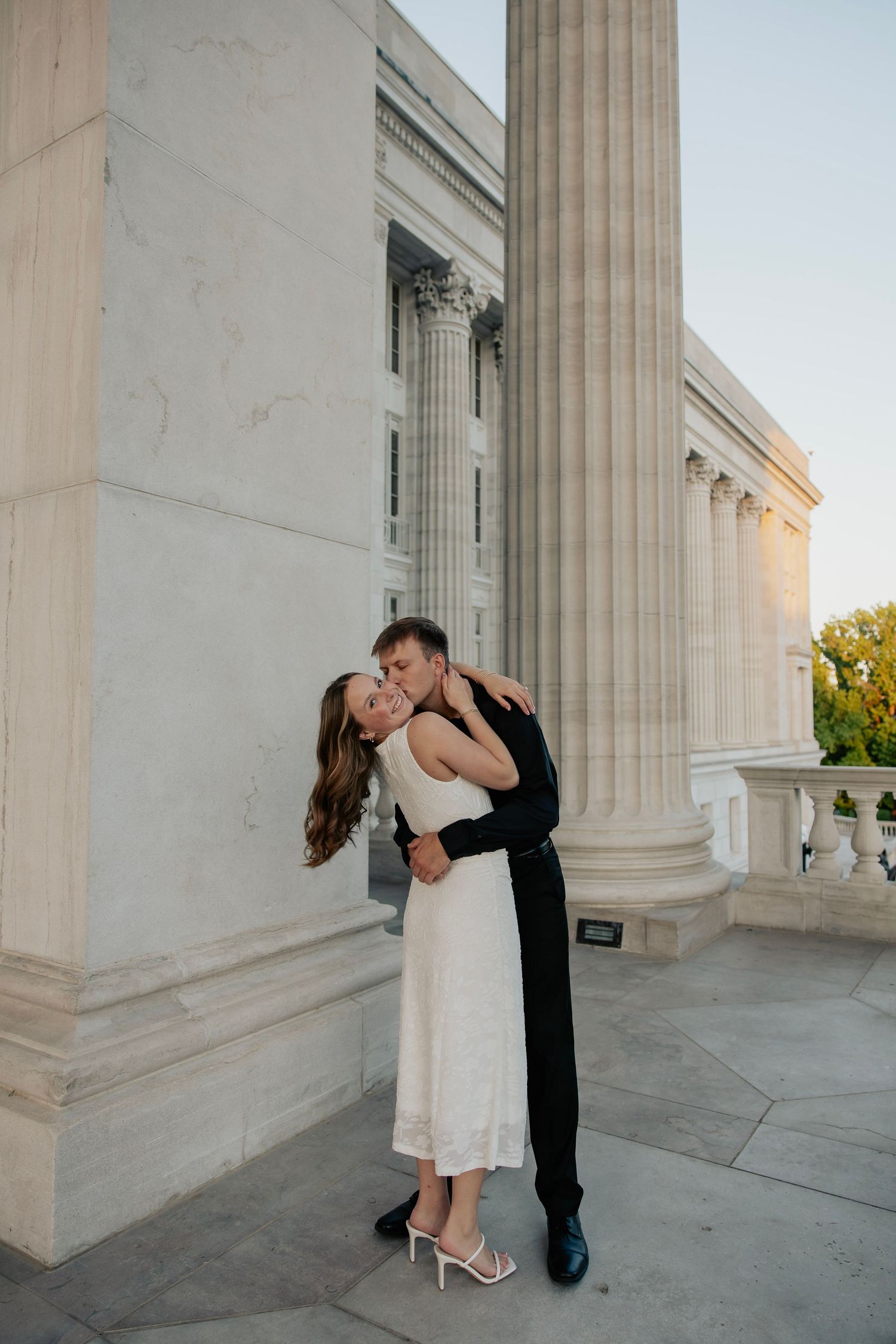 Groom dipping and kissing bride in white dress beside tall marble columns at sunset.
