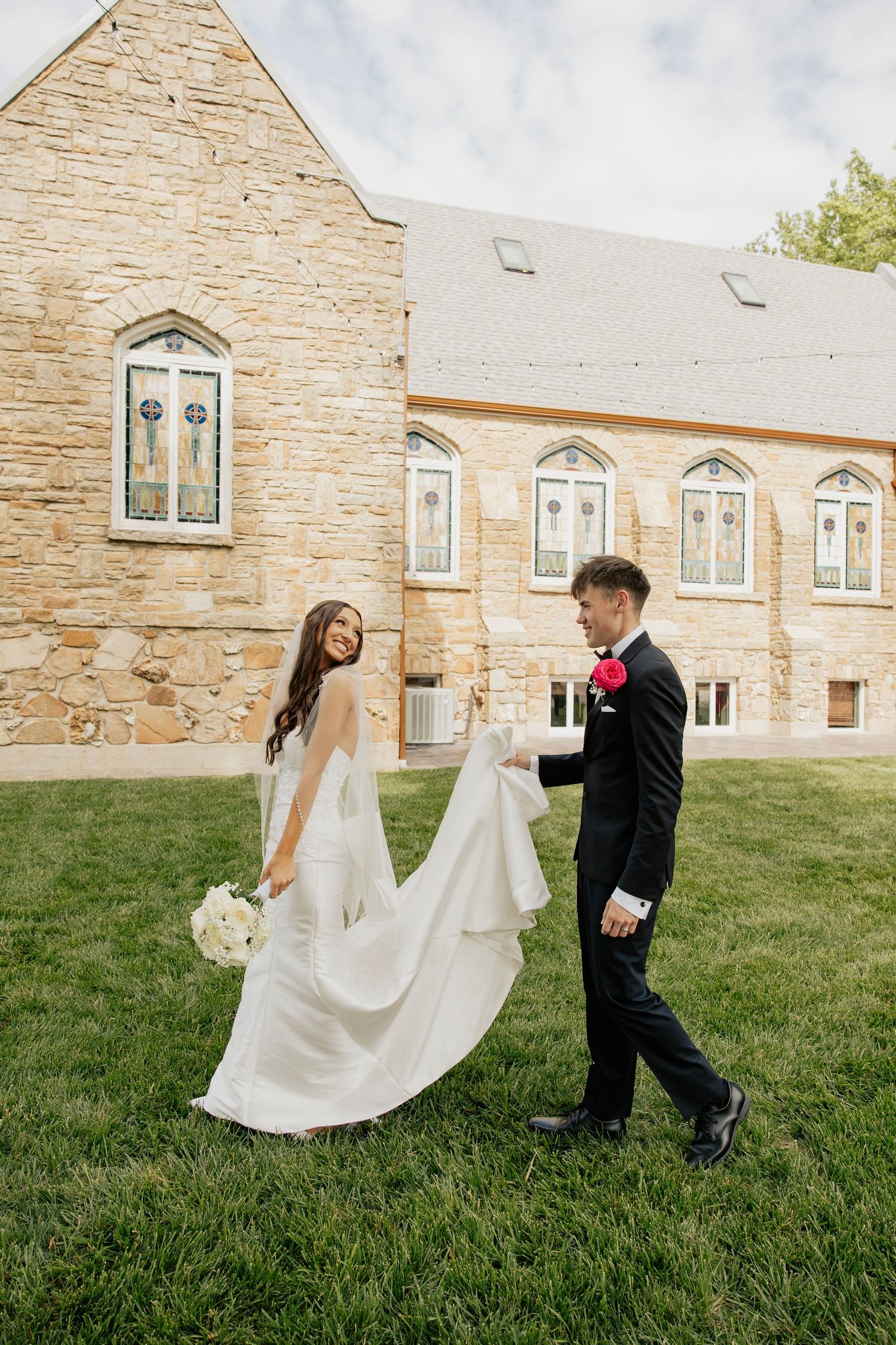 Smiling bride in a white gown walks with groom outside a stone church with stained glass windows.