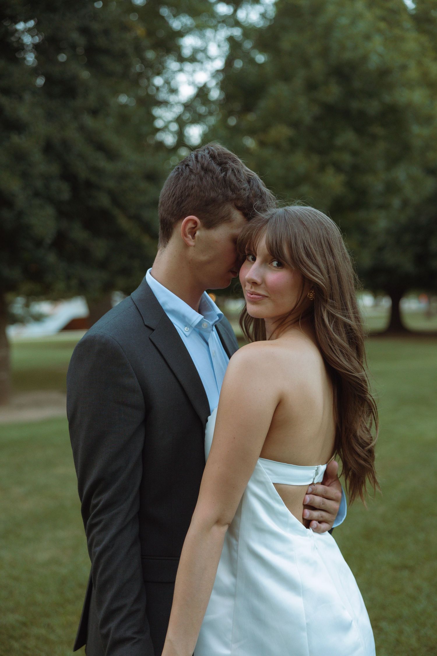 A couple embraces outdoors, man in dark suit kissing woman's head as she looks back in a white strapless dress.