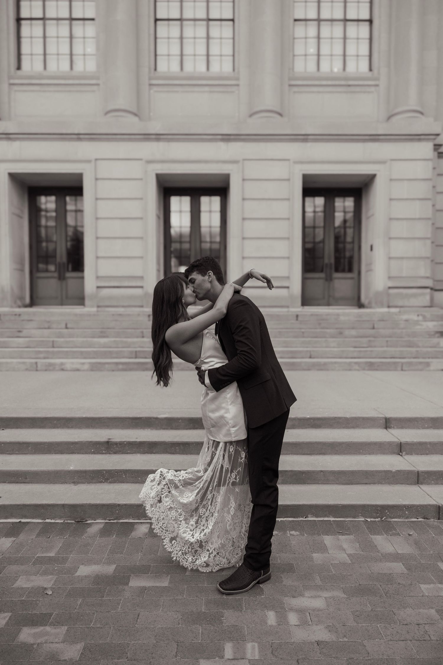 Black and white photo of a couple sharing a romantic kiss in wedding attire on steps of a classic building.