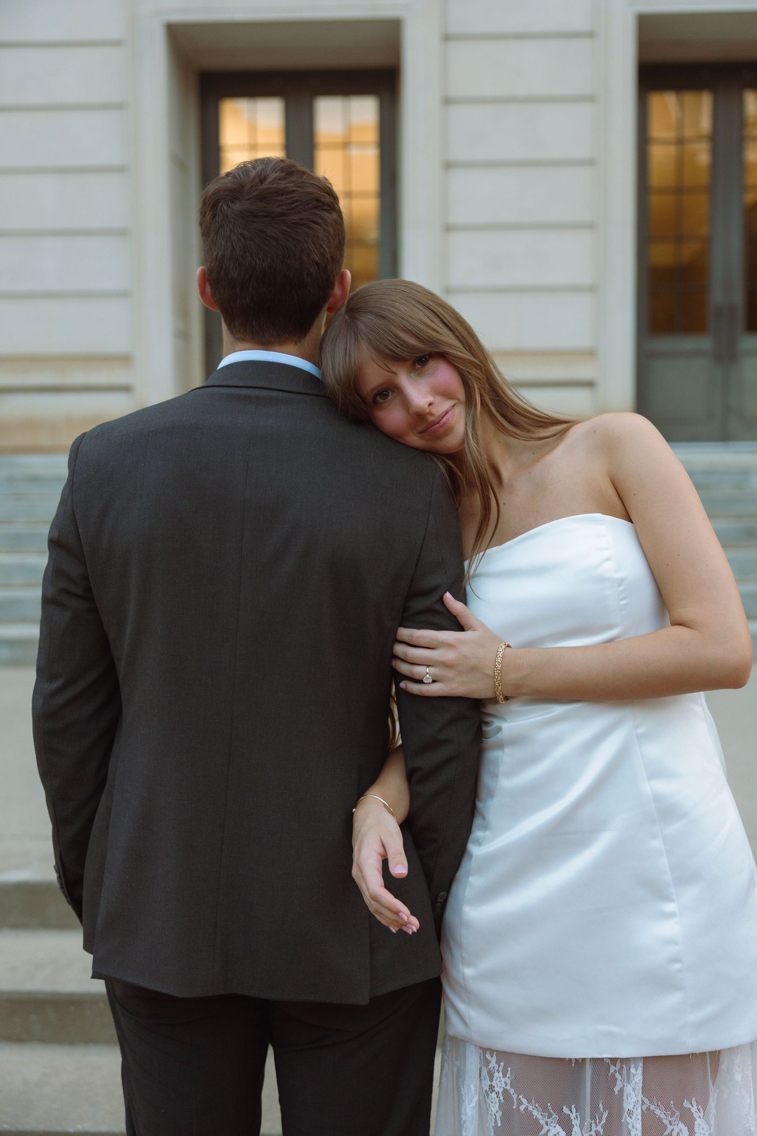 Bride in white strapless dress rests head on groom's shoulder in black suit on steps.