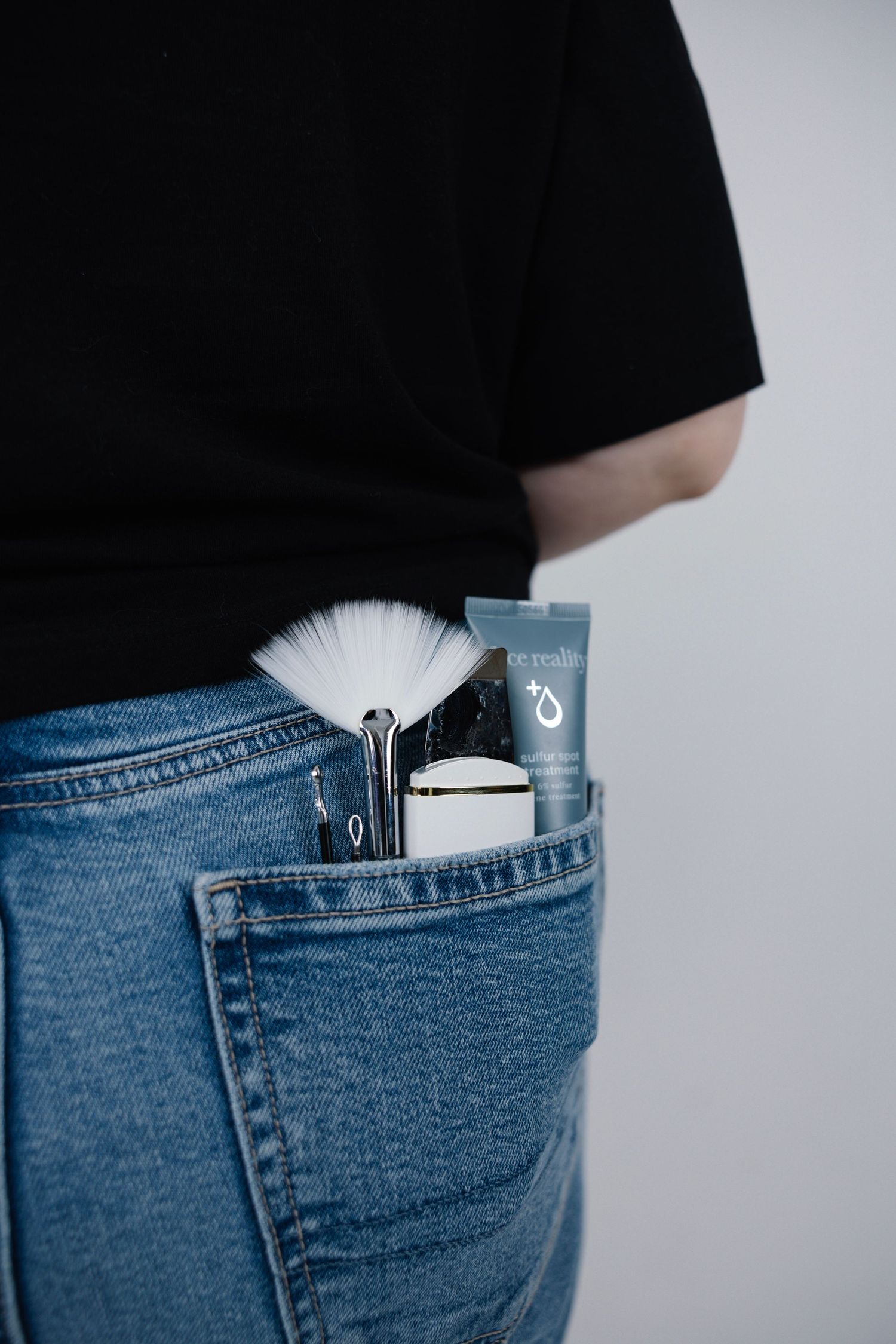 Makeup brushes, skincare tube, and beauty tools tucked into the back pocket of blue jeans.