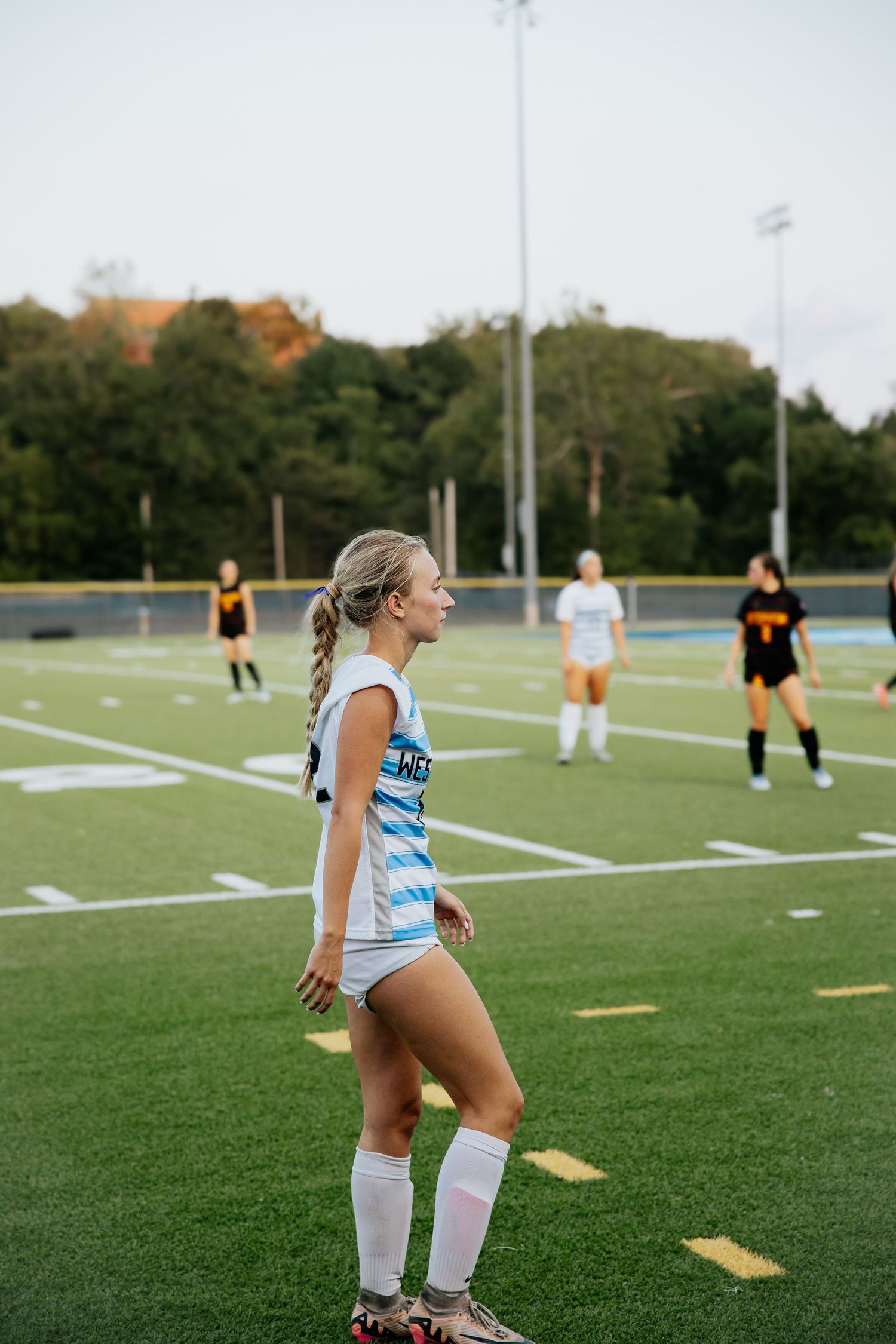 Female soccer player in white uniform stands on green turf field during evening game with teammates.