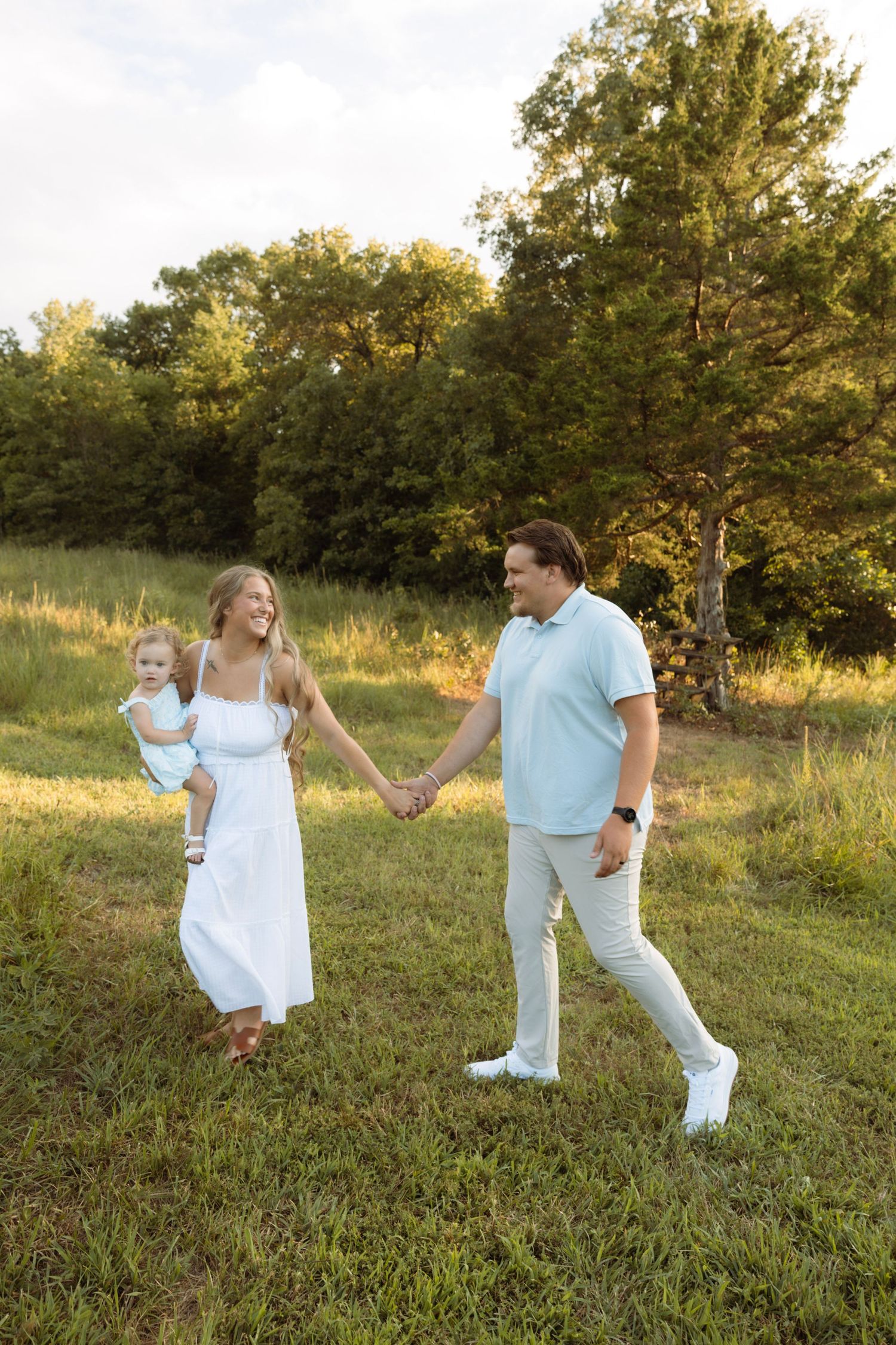 A happy family walks hand-in-hand through a green meadow, mother in white dress holding baby, father in light blue polo.